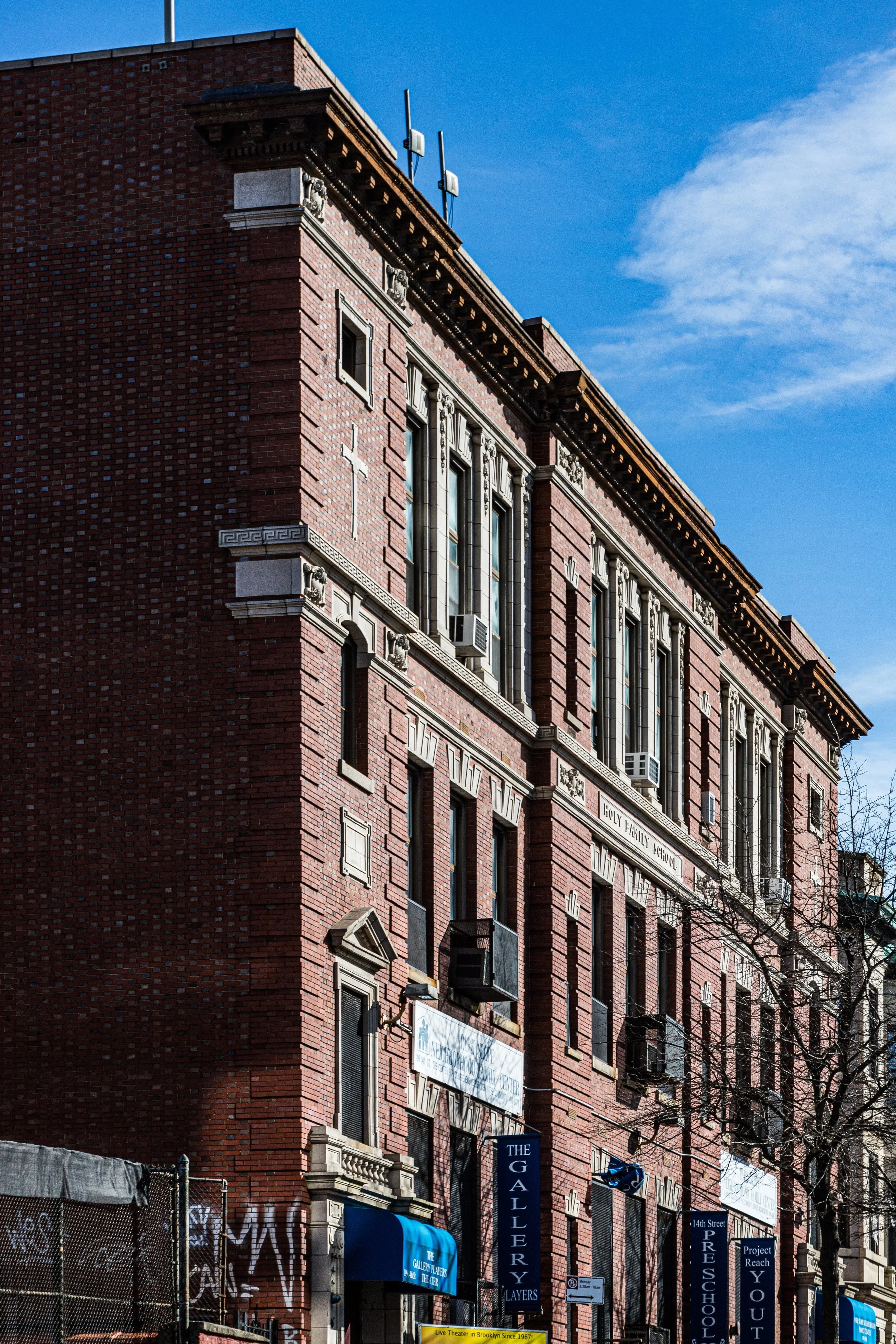 A red brick building with decorative stone accents and tall windows, some with air conditioning units. There are blue signs at the bottom for galleries, preschool, and outreach services. The sky is clear with some clouds.