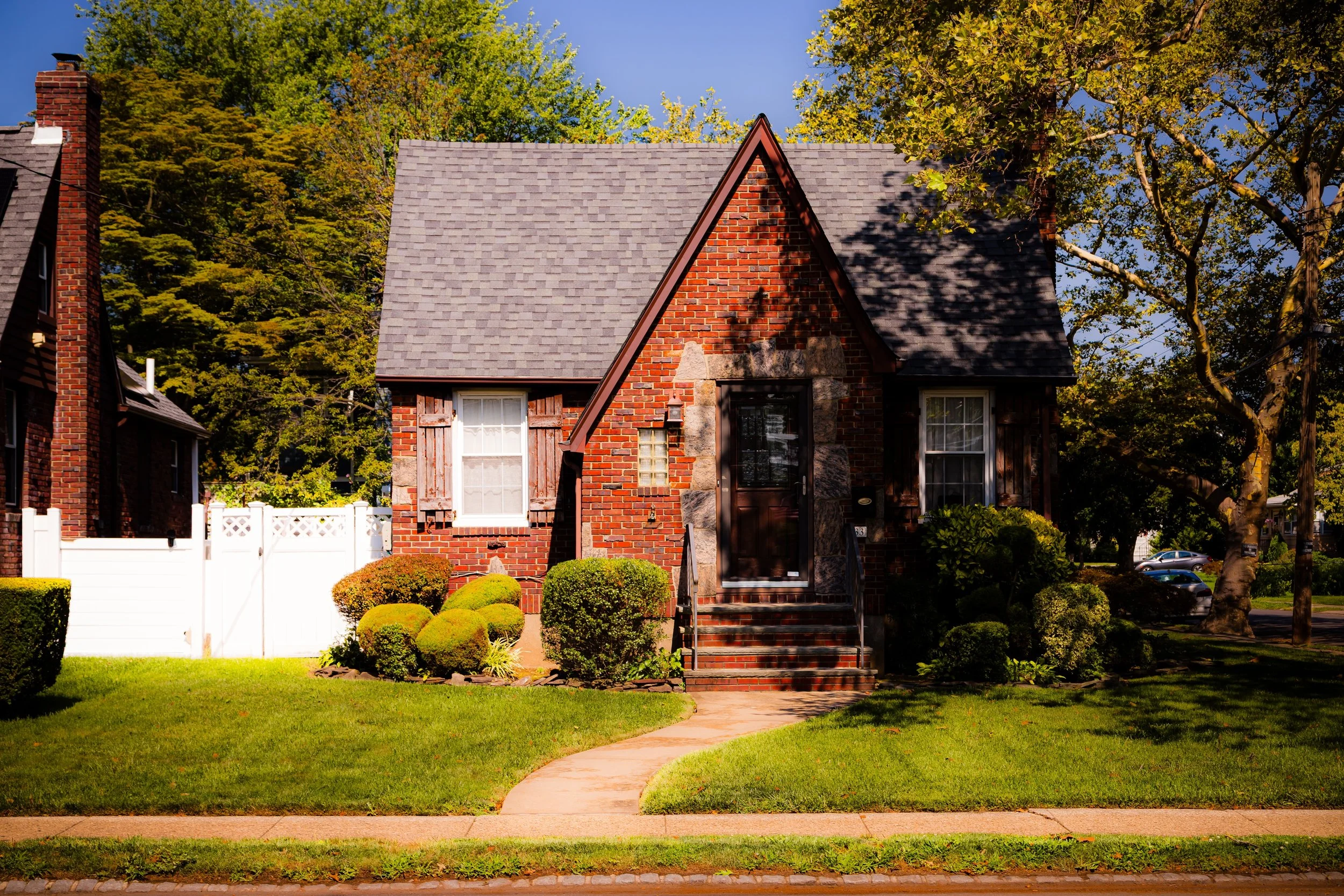 A small brick house with a gabled roof, two windows with wooden shutters, and a front door accessible by brick stairs. The house is surrounded by a well-manicured lawn and bushes, with a curved sidewalk leading up to the entrance. Trees with green leaves are in the background, and a white fence is visible on the left side.