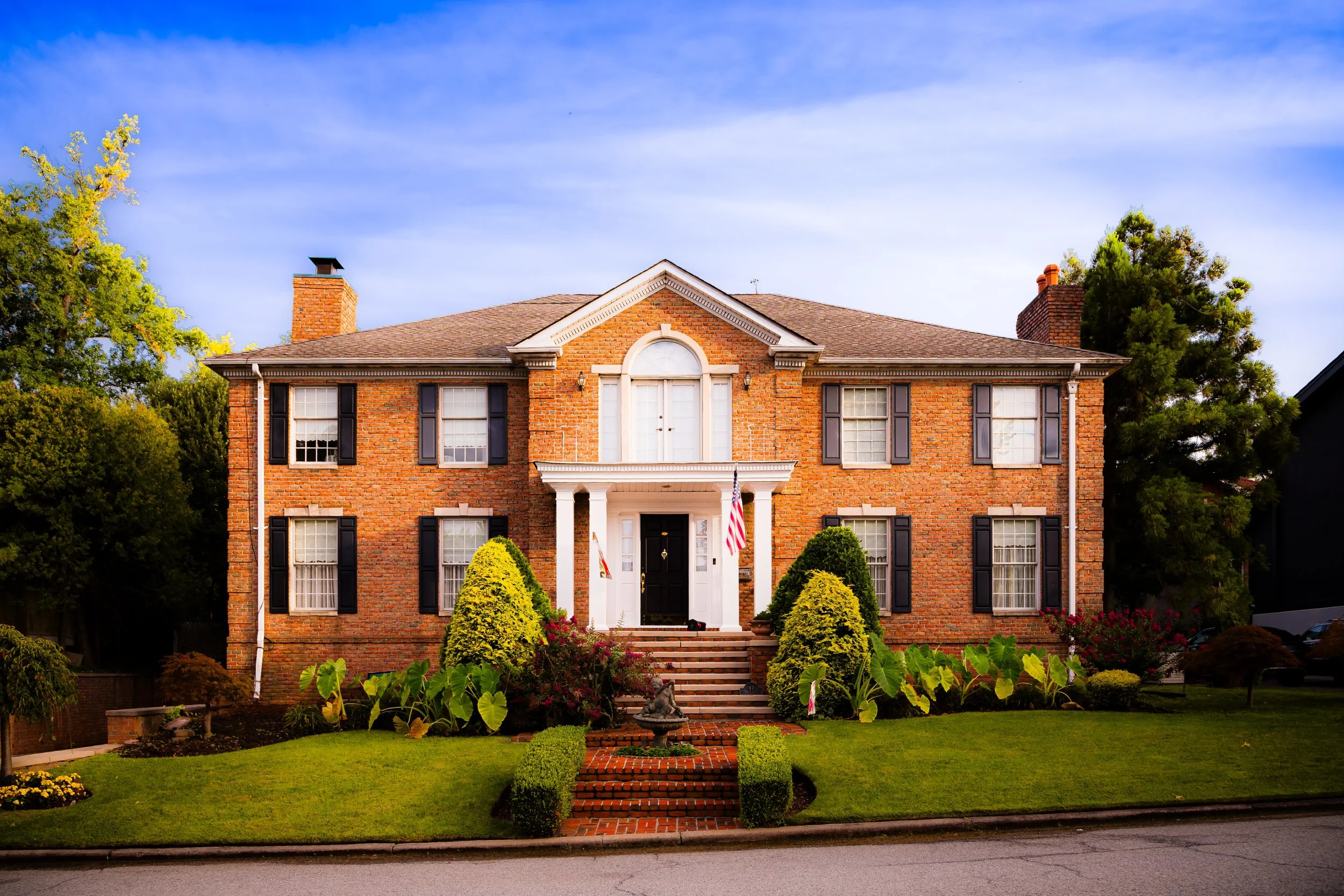 A large two-story brick house with a front porch, white columns, and steps leading down to a well-manicured lawn with shrubs and flowers, under a blue sky.