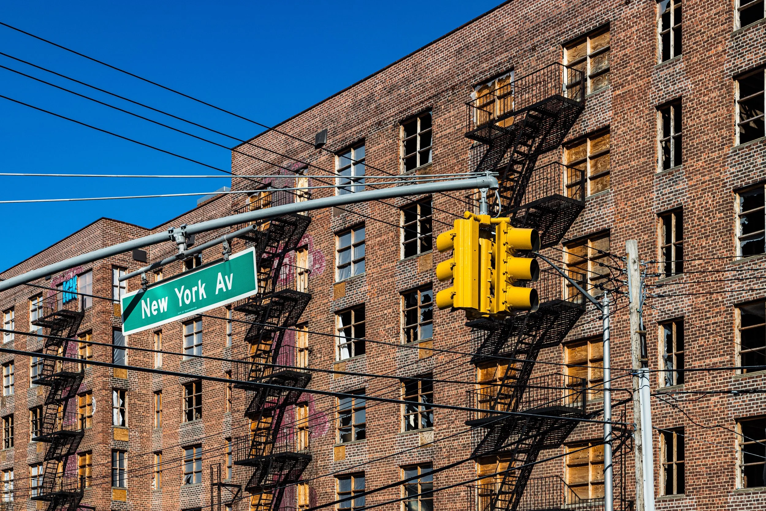 A brick apartment building with broken and boarded-up windows, black fire escapes, and a bright yellow traffic light, with a street sign reading 'New York Av' and power lines overhead.