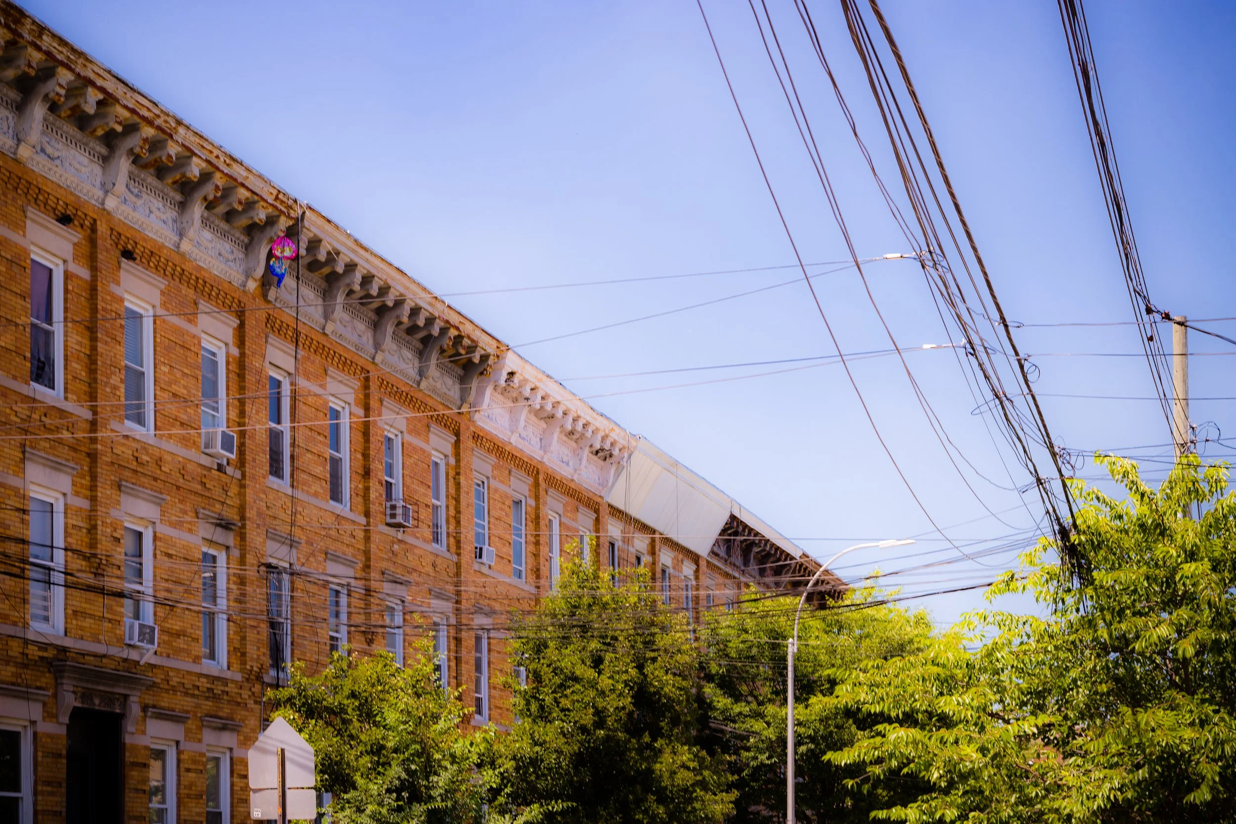 An urban scene with a brick building with multiple windows, some with air conditioning units, trees in the foreground, and overhead utility wires against a blue sky.