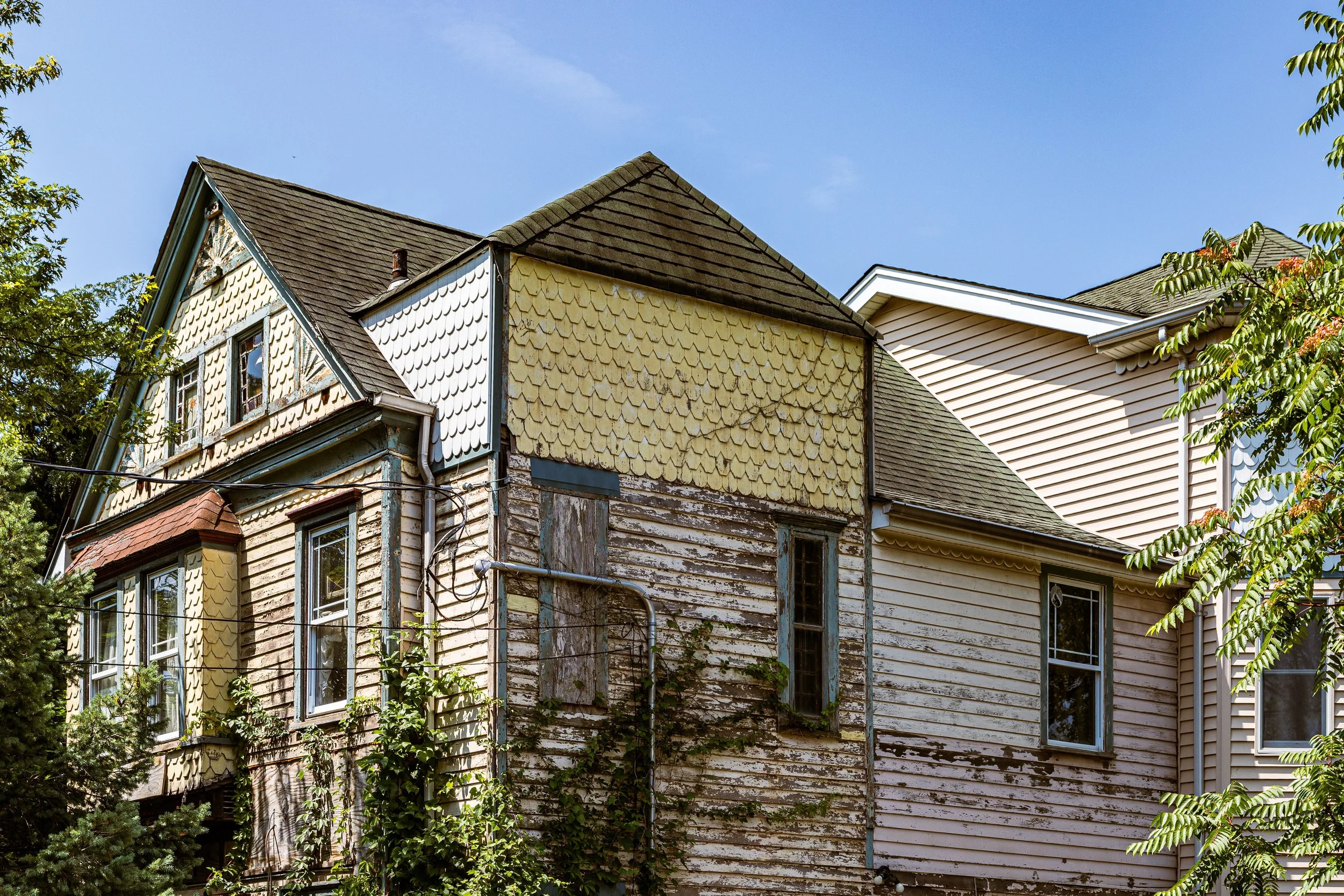 Old, weathered house with peeling paint and mixed siding, neighboring a newer, well-maintained house, under clear blue sky.