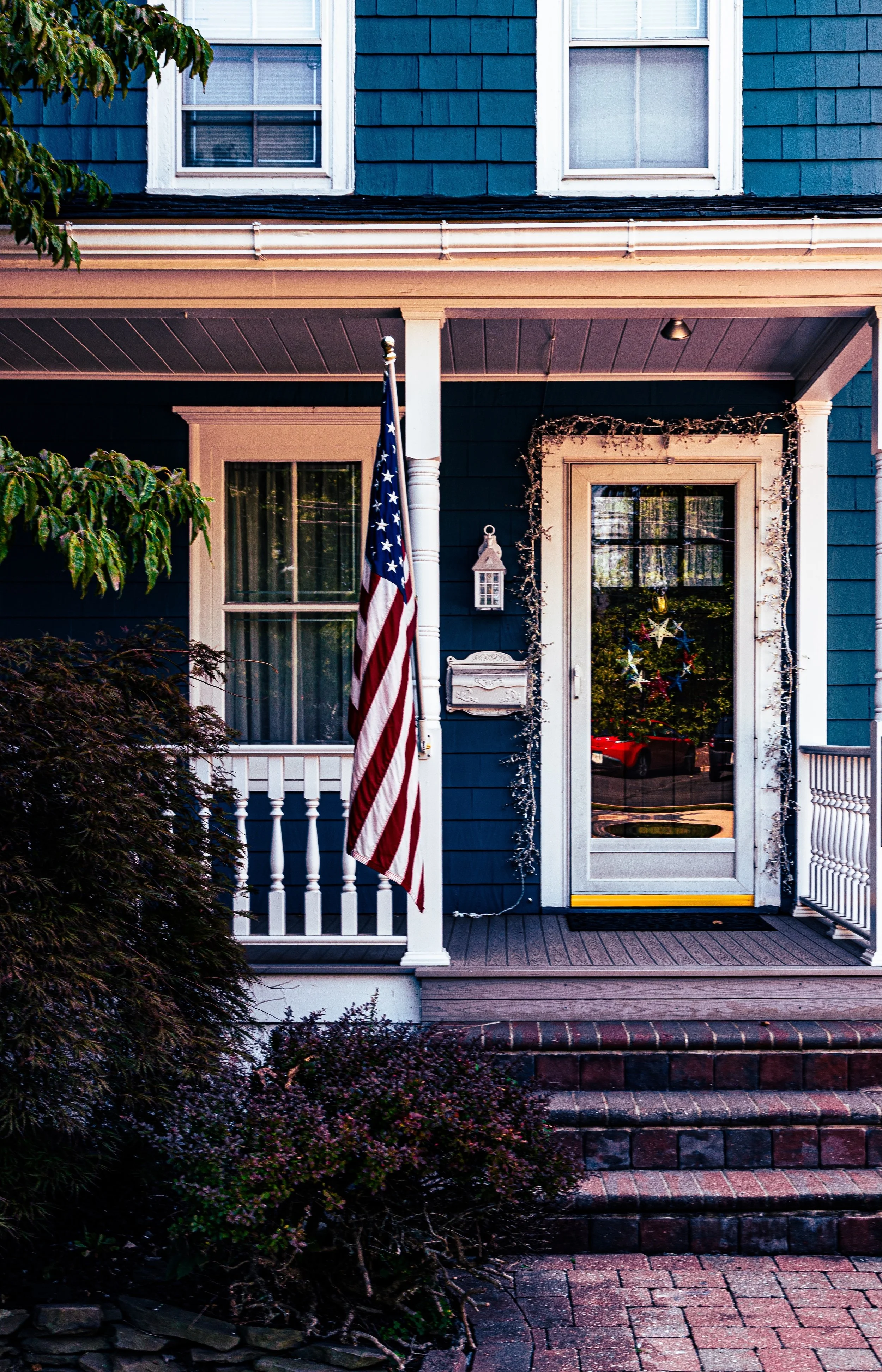 The front porch of a blue house decorated with a Christmas wreath, string lights, and an American flag.