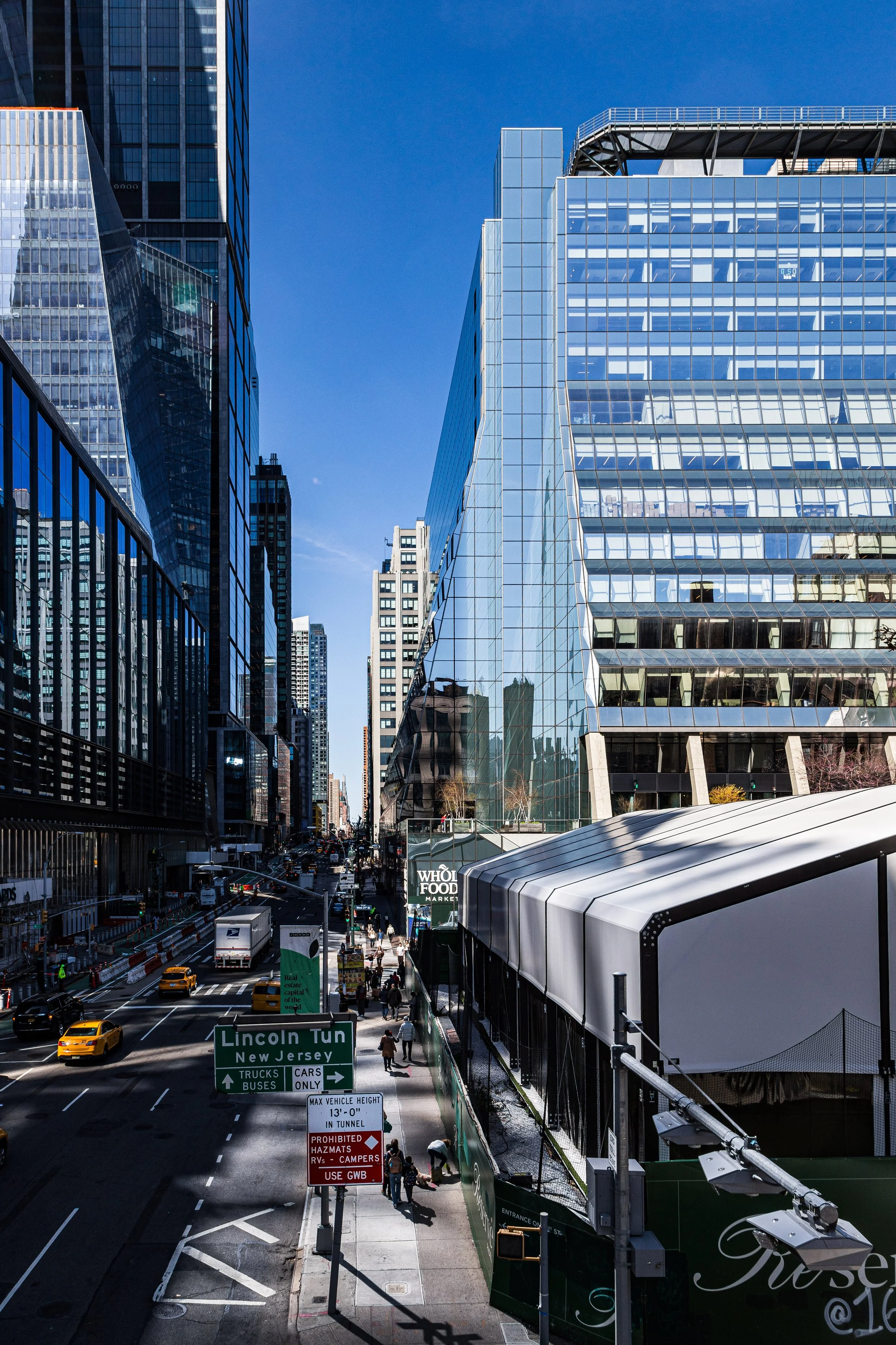 A city street in New York City with tall glass skyscrapers, yellow taxis, and people walking. Signs indicate Lincoln Tunnel and rules for vehicles and pedestrians.