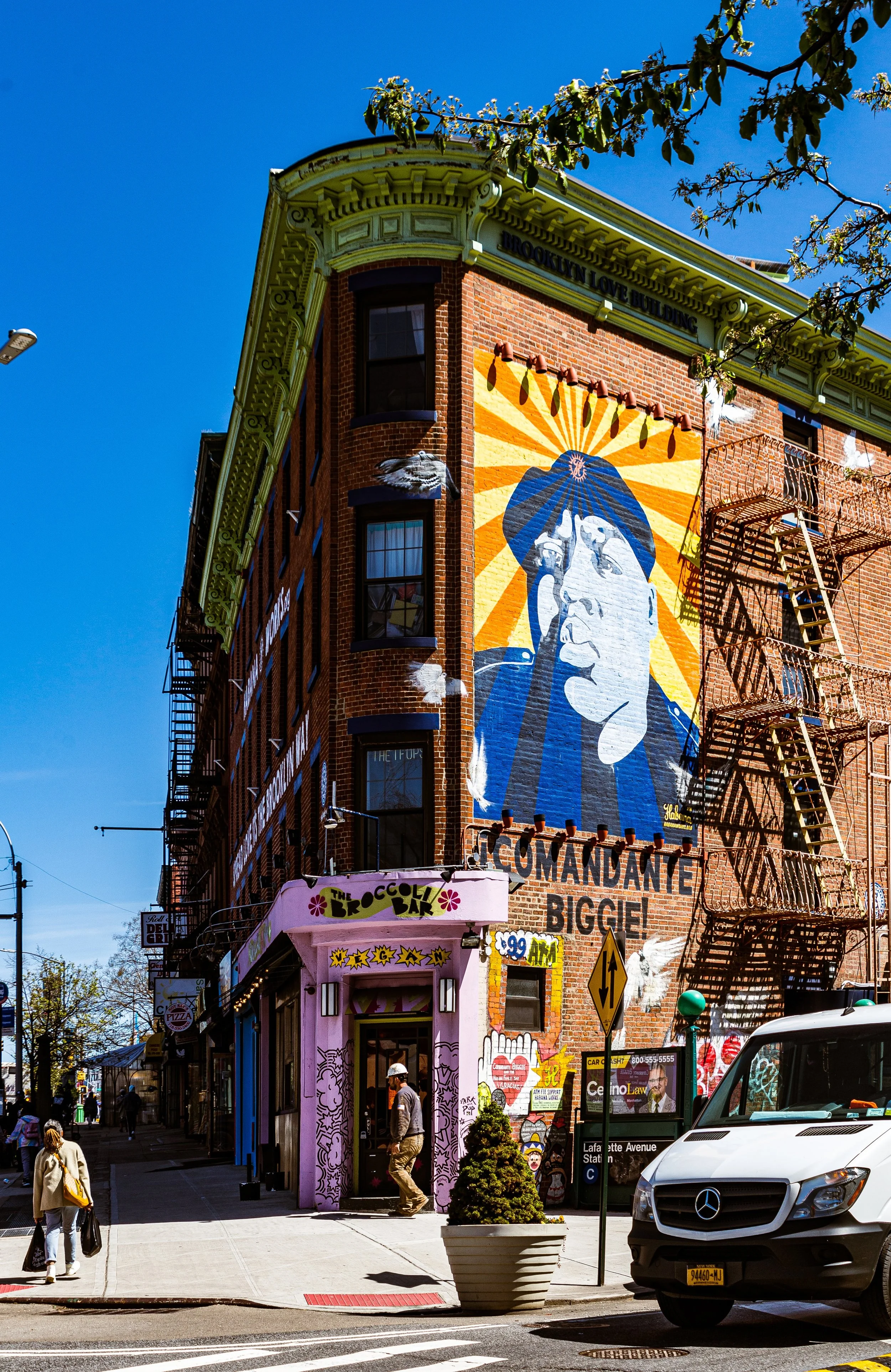 Street view of a corner building with a large mural of a woman holding a phone on the side wall, fire escape stairs, and a pink storefront called 'The Brooklyn Bar', with pedestrians and a white van in the foreground.