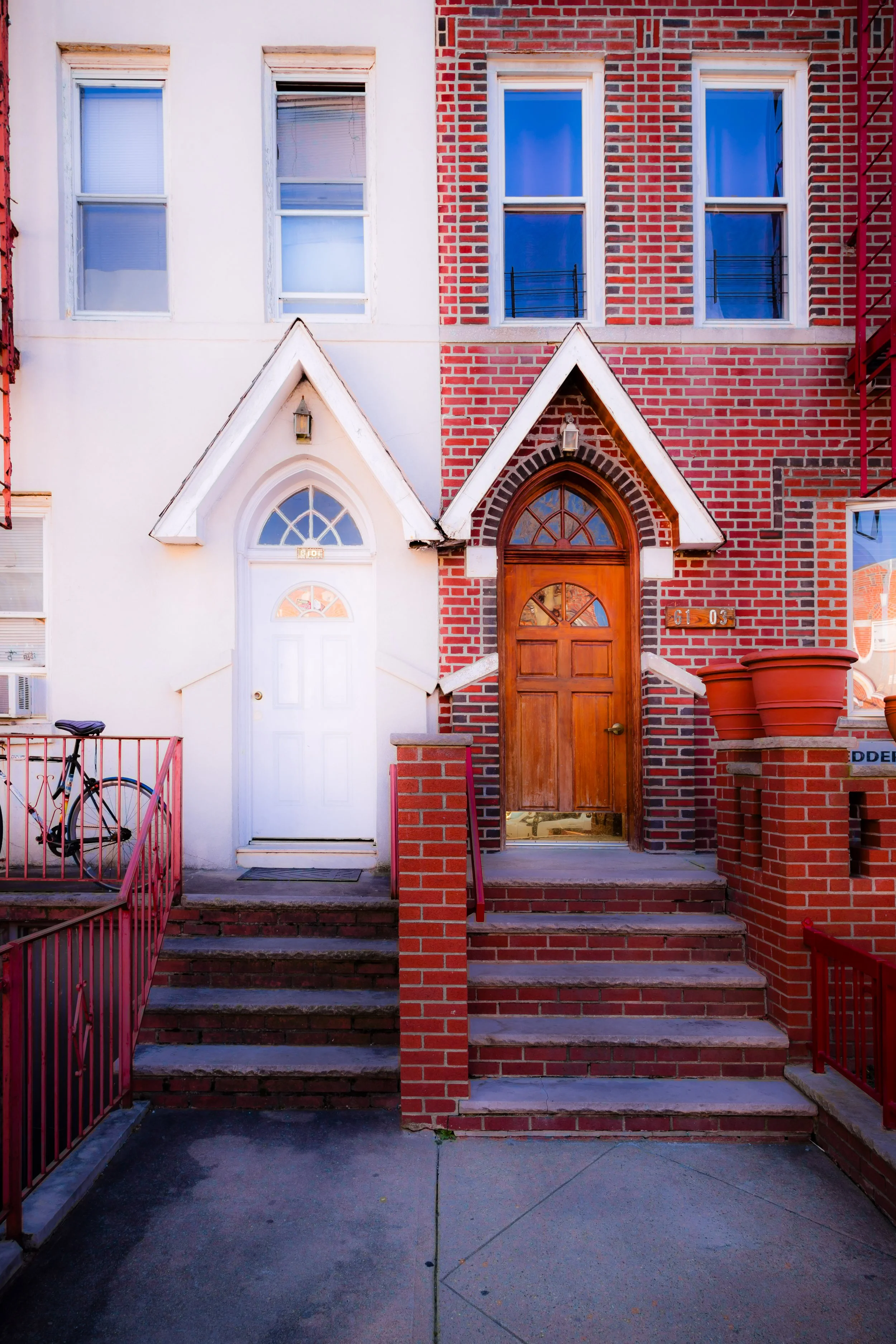 View of two adjacent houses with front steps, red brick, and doors; one house has a white door, the other has a wooden door; bike parked on the stairs railing.