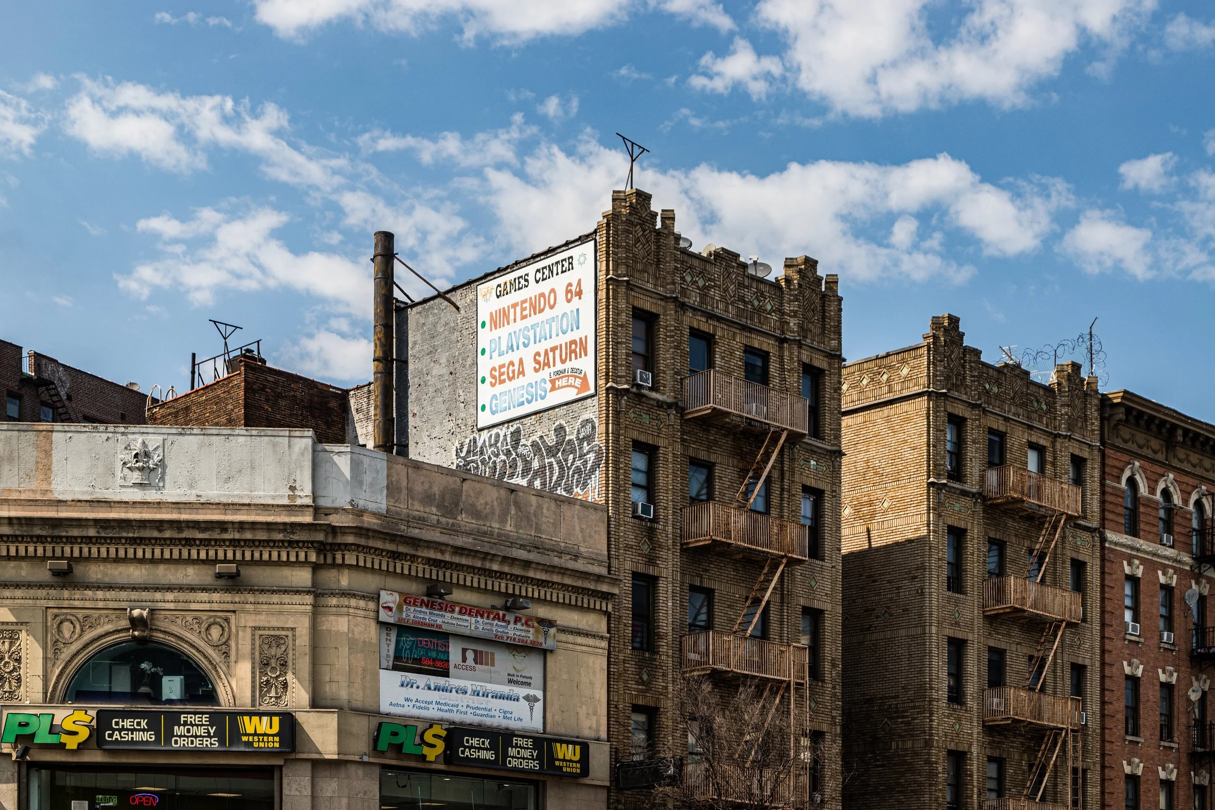Tall brick buildings with fire escapes in an urban setting under a partly cloudy sky, with a large billboard advertising gaming consoles like Nintendo 64, PlayStation, Sega Saturn, and Genesis.