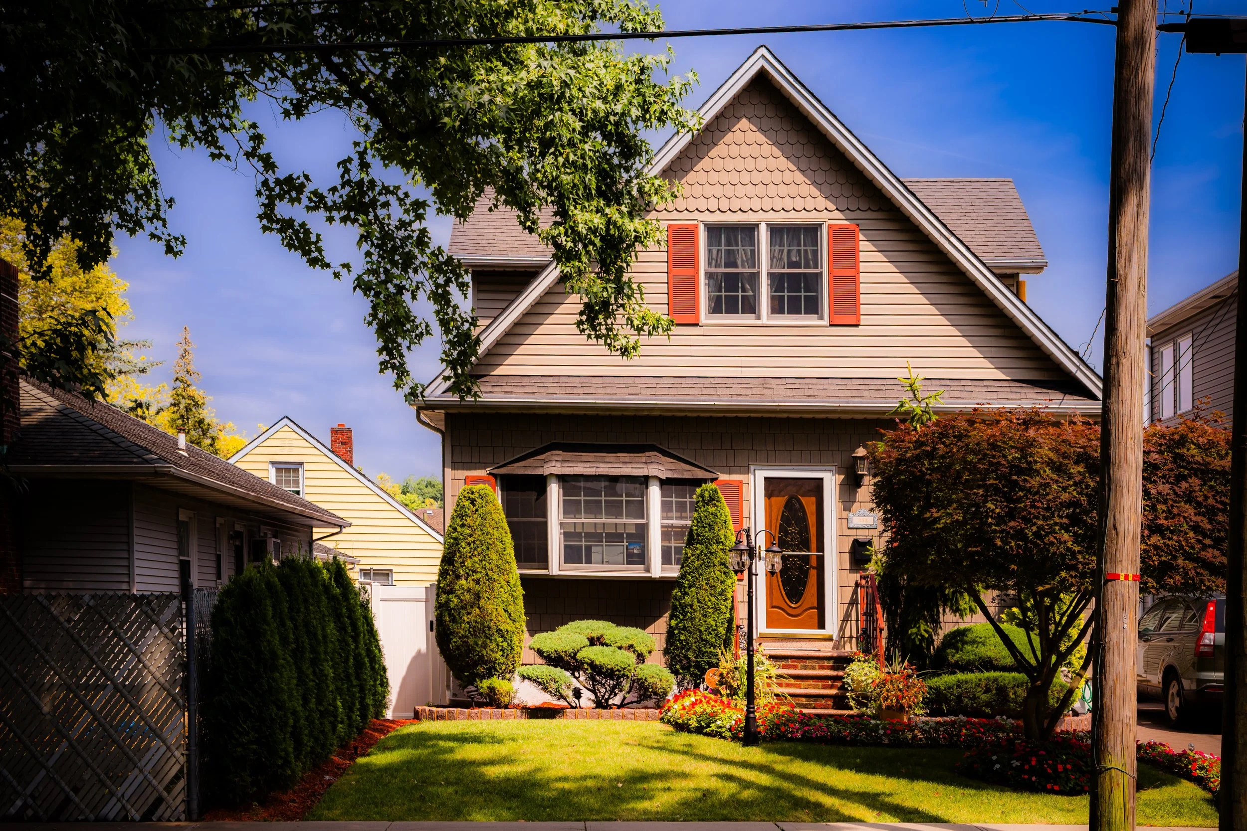 A two-story house with tan siding, red shutters, a small front porch with stairs, green lawn, and landscaped garden with bushes and flowers, under a blue sky with some clouds.