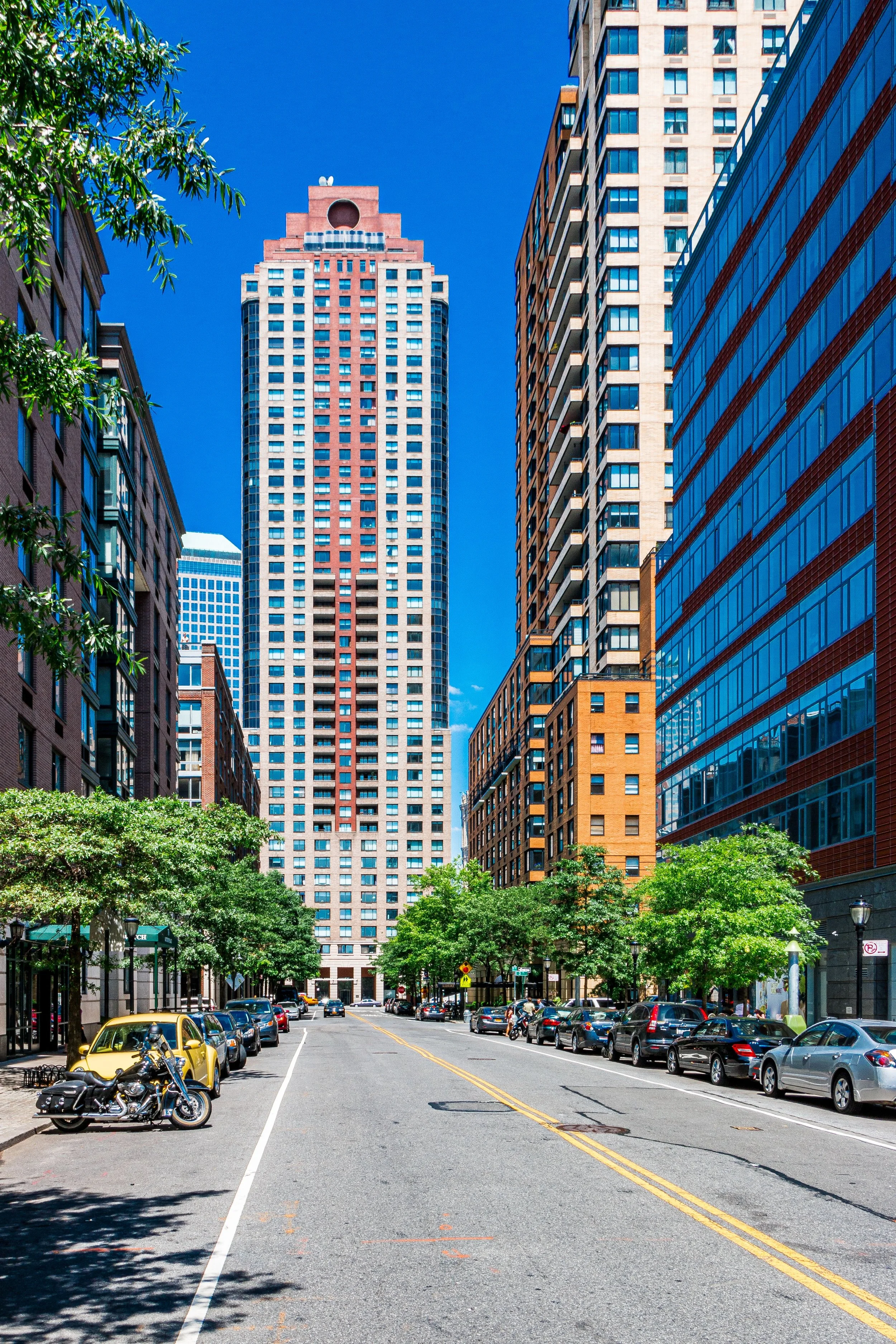 A city street lined with trees, parked cars, and a motorcycle, leading towards tall modern buildings under a bright blue sky.