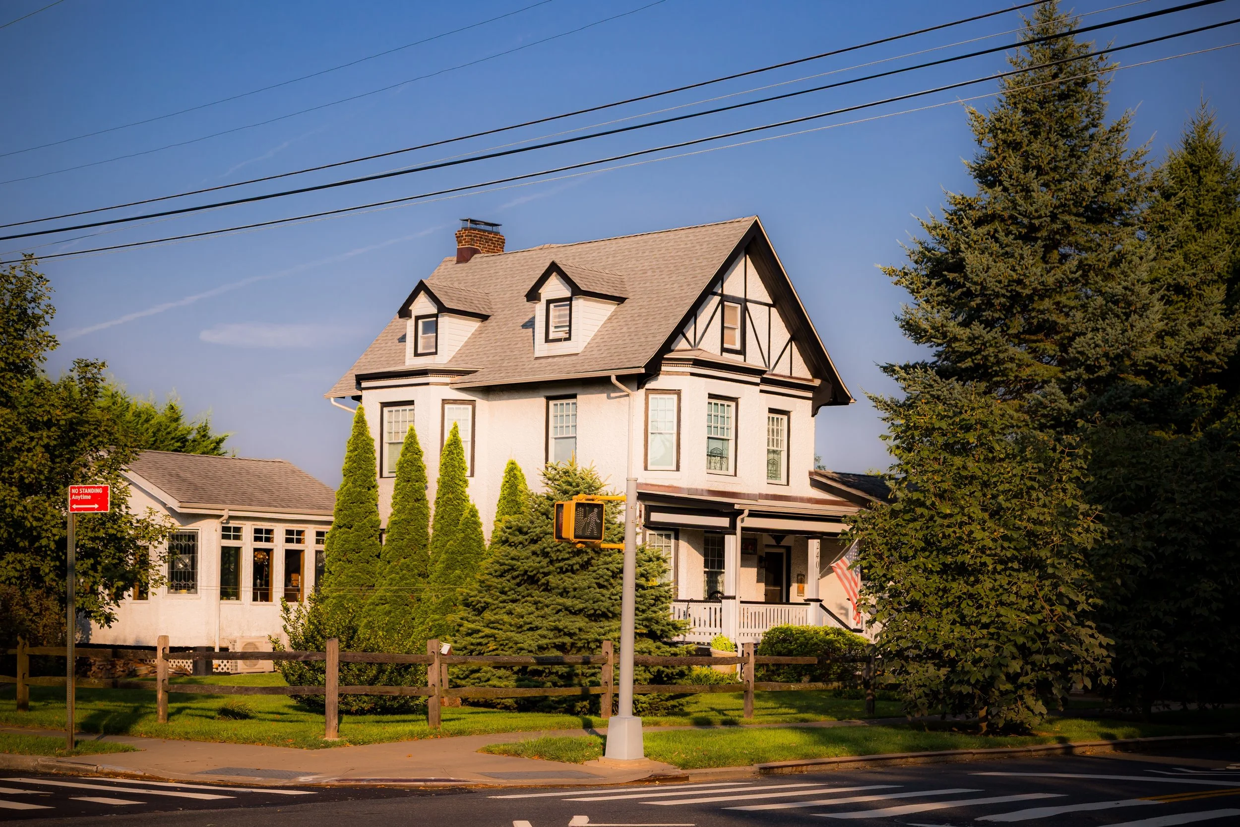 A large two-story house with a beige exterior, dark trim, and a steep roof with dormer windows. The house is surrounded by tall trees, a small front lawn, and a wooden fence. A sidewalk and street are visible in the foreground, with a traffic signal and power lines overhead.