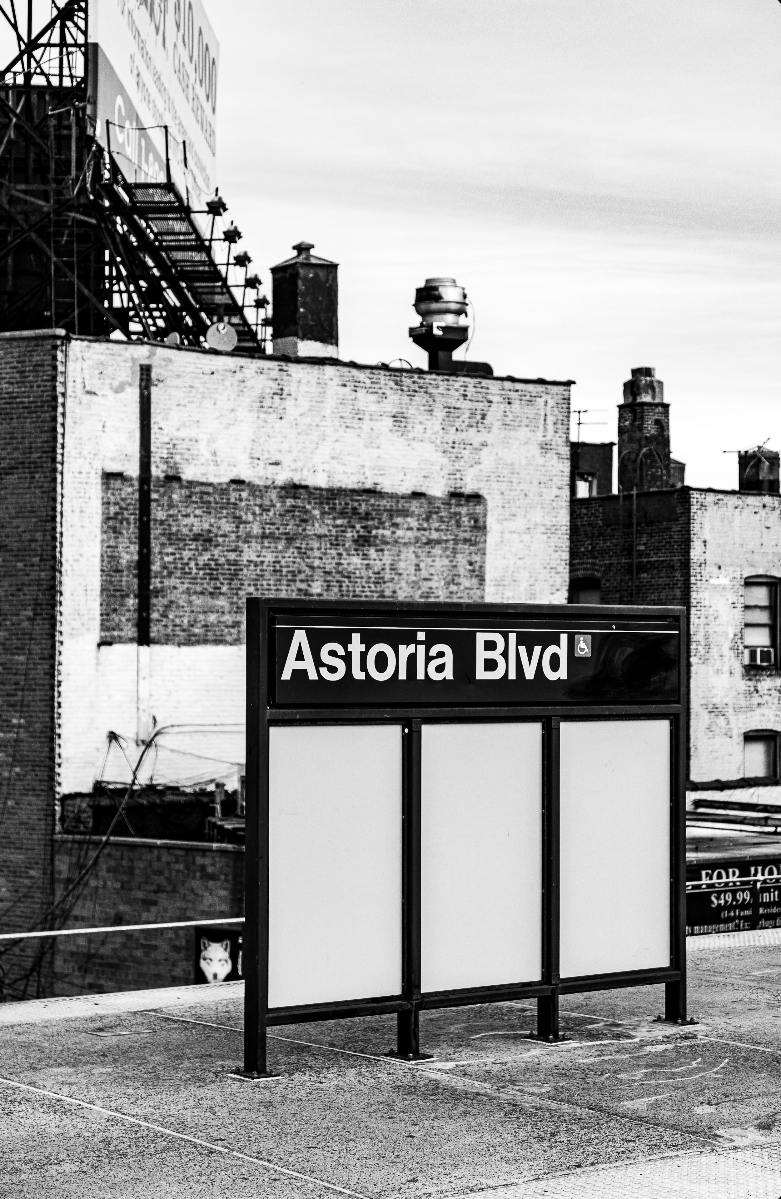 Black-and-white image of a city street sign reading 'Astoria Blvd,' with a blank billboard underneath, surrounded by urban buildings and rooftops.