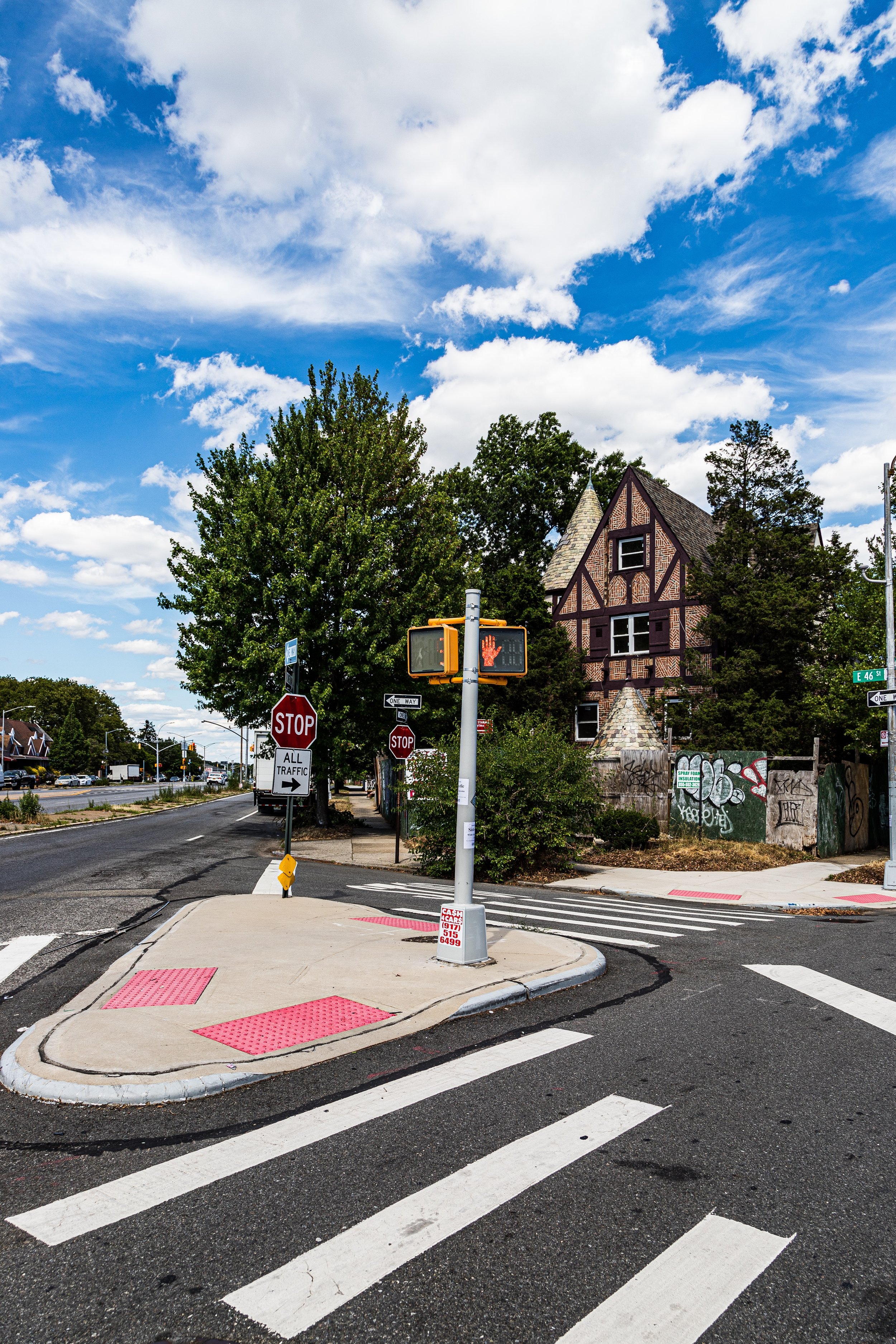 A city crosswalk with a pedestrian signal showing a hand, stop signs, and a corner of a street with a brick house in the background under a partly cloudy blue sky.
