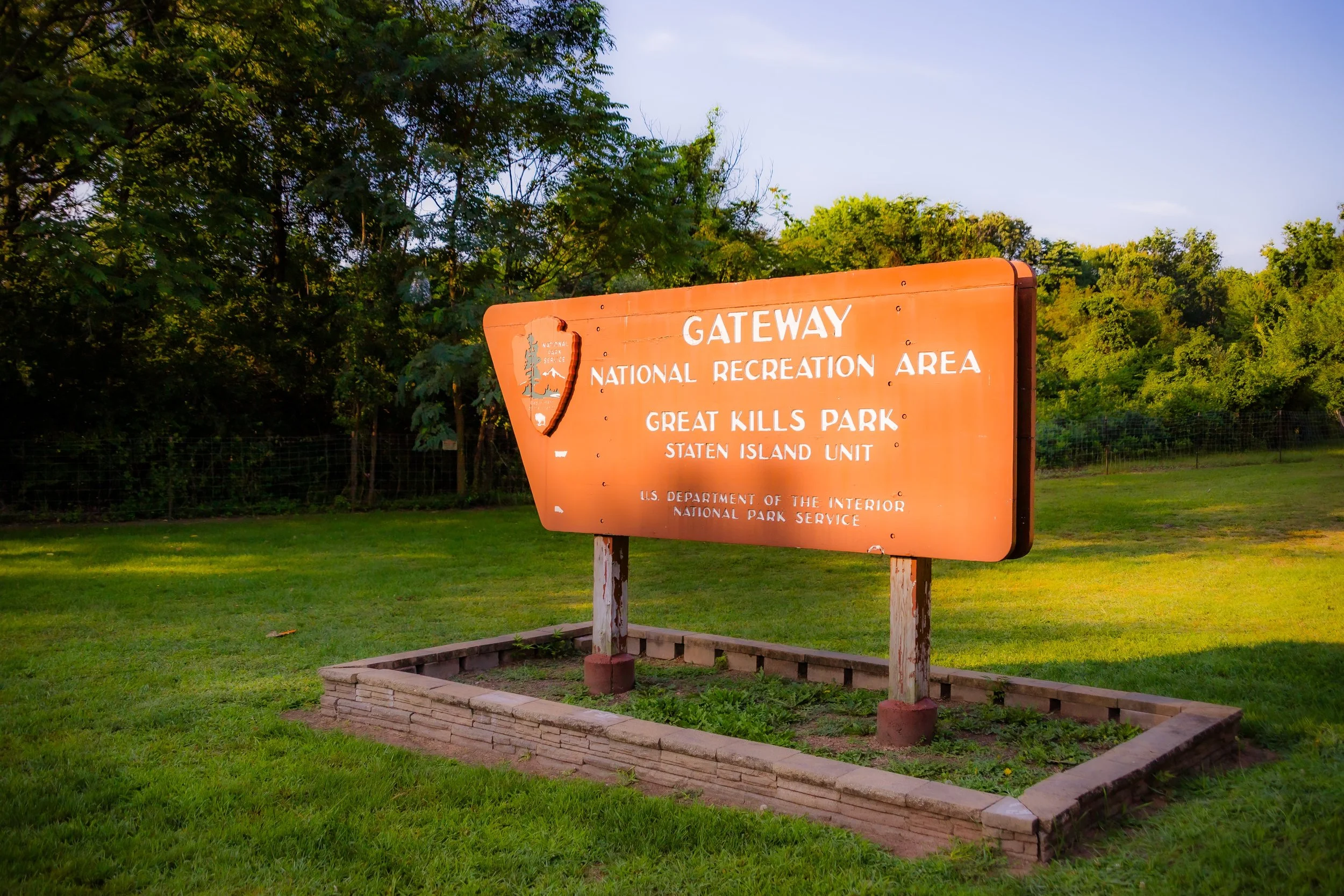 A large orange sign indicating the entry to the Gateway National Recreation Area at Great Kills Park, Staten Island Unit, with the National Park Service emblem and information, set in a grassy area with trees in the background.