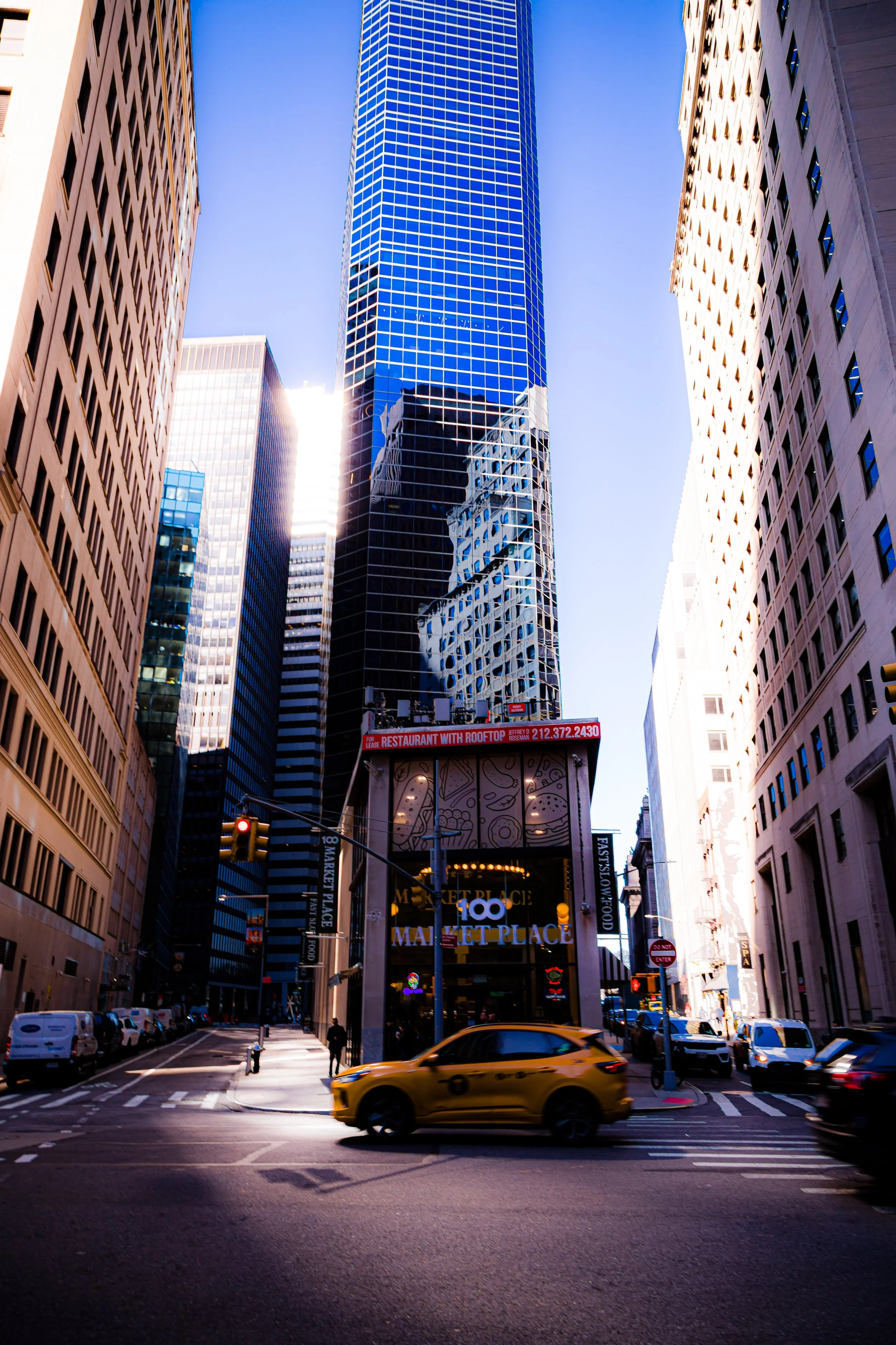 Tall glass skyscraper reflecting nearby buildings, with busy city street below showing cars and pedestrians at intersection.