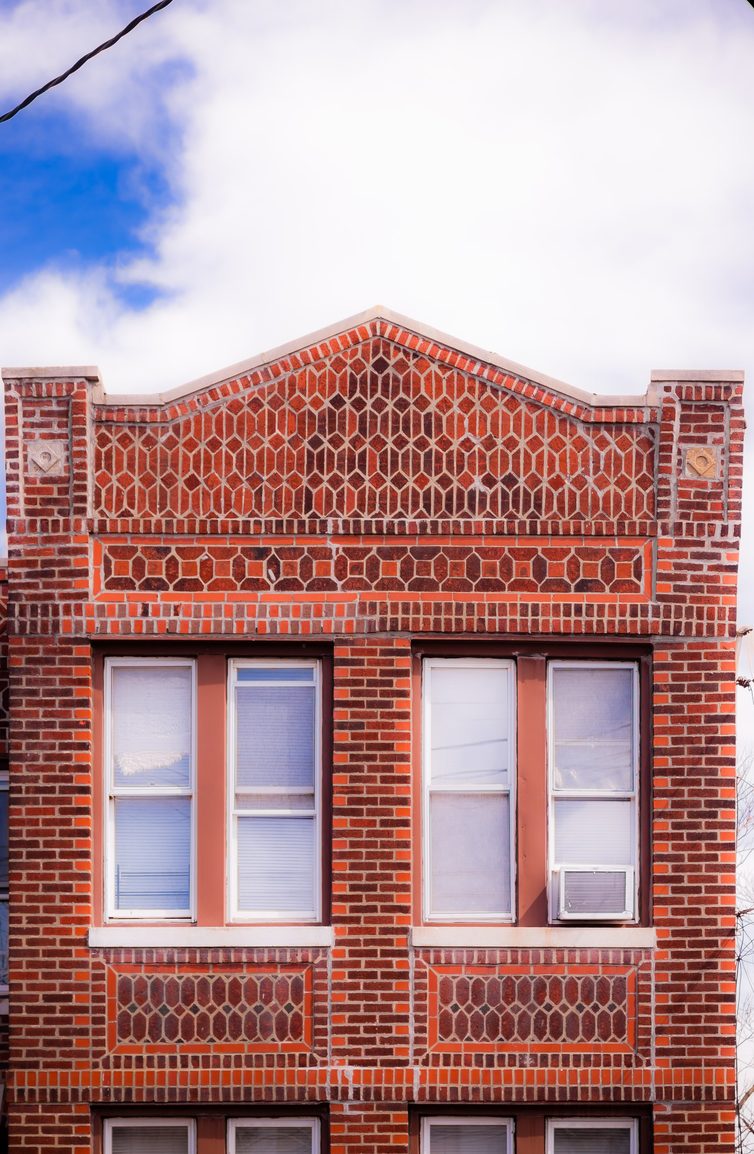 Close-up of an old brick building facade with patterned brickwork above and below windows with blinds, set against a partly cloudy sky.