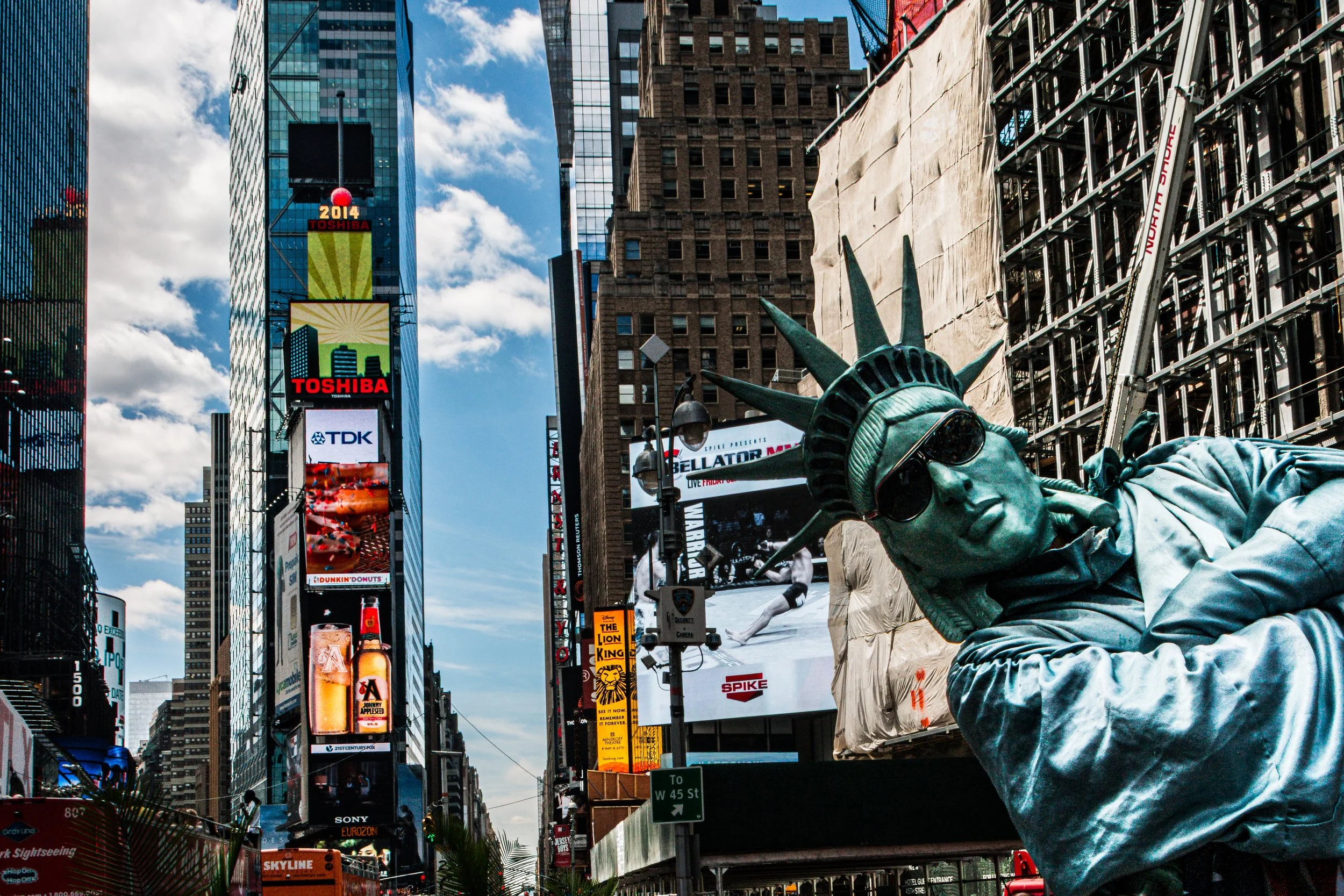 Statue of Liberty replica wearing sunglasses in Times Square, New York City, with tall buildings and electronic billboards in the background.