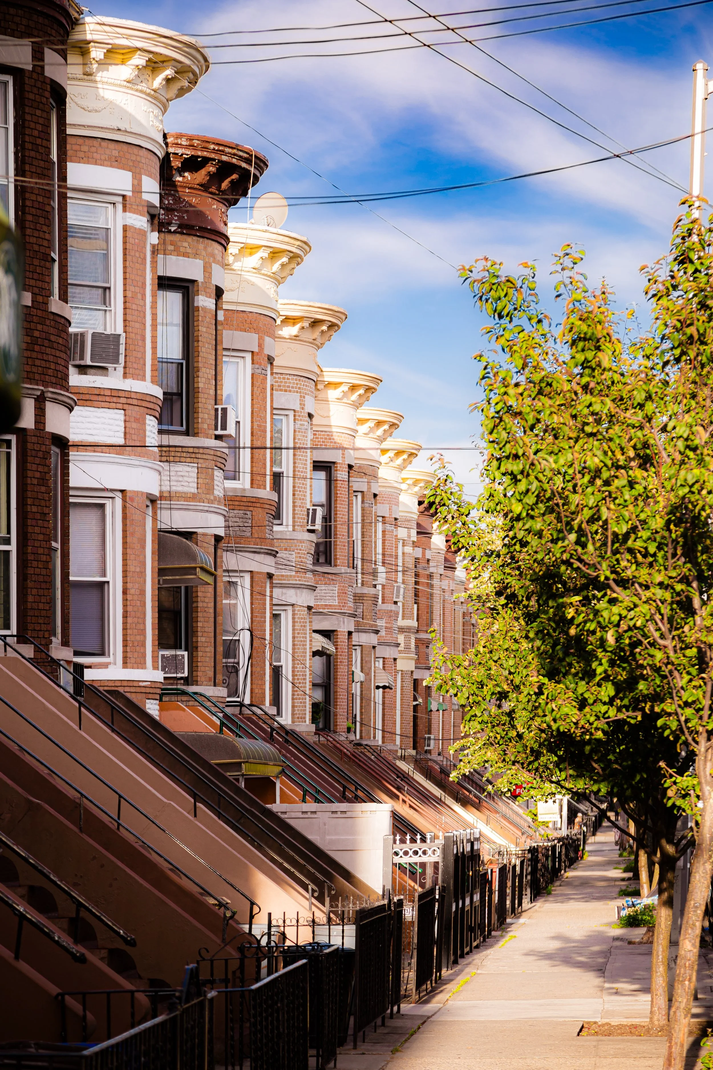 Row of brownstone brown brick townhouses with stoops and black iron fences along a sunny sidewalk, with trees and a blue sky overhead.