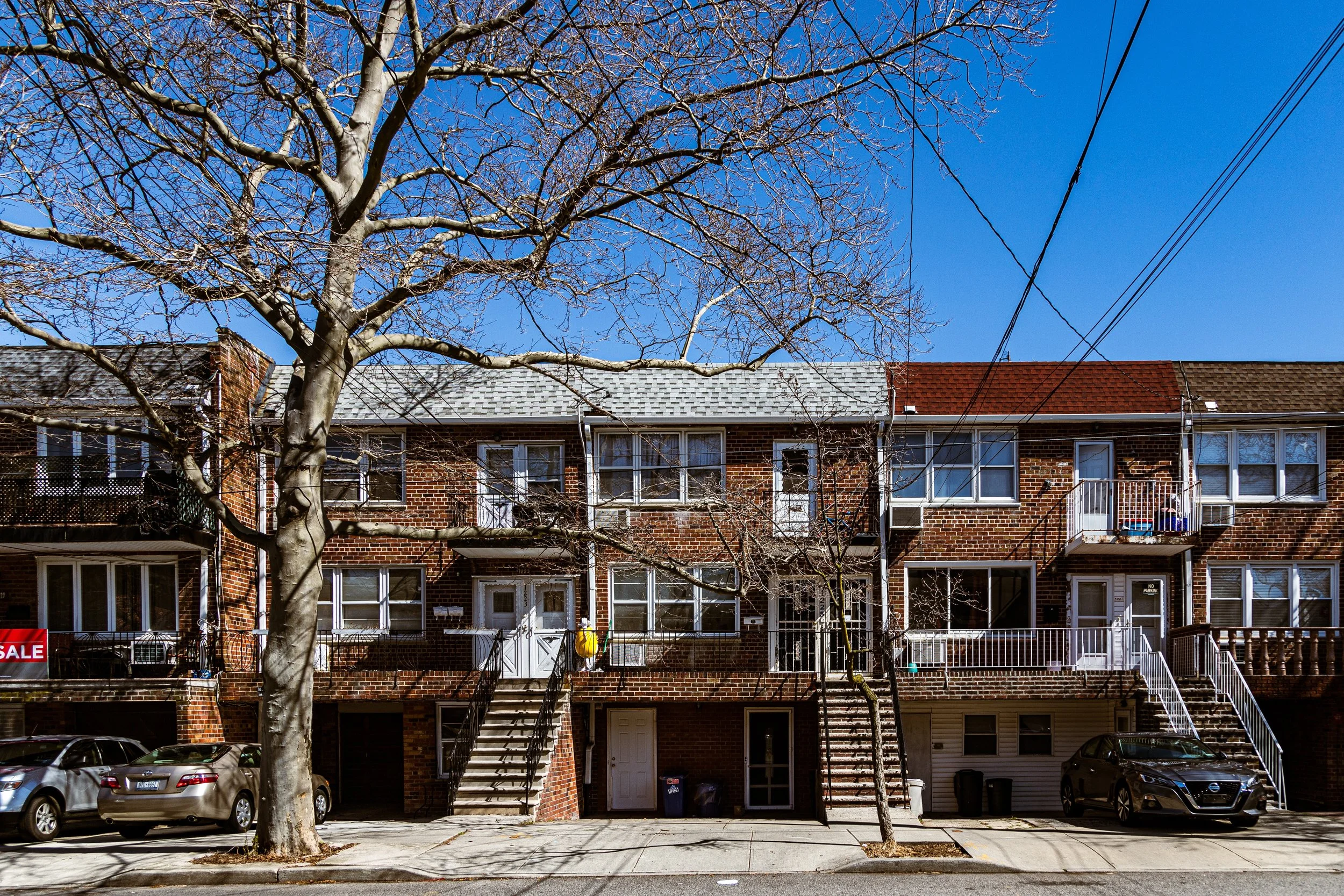 Brick row of residential apartments with exterior stairs, parked cars, leafless trees, and a clear blue sky.