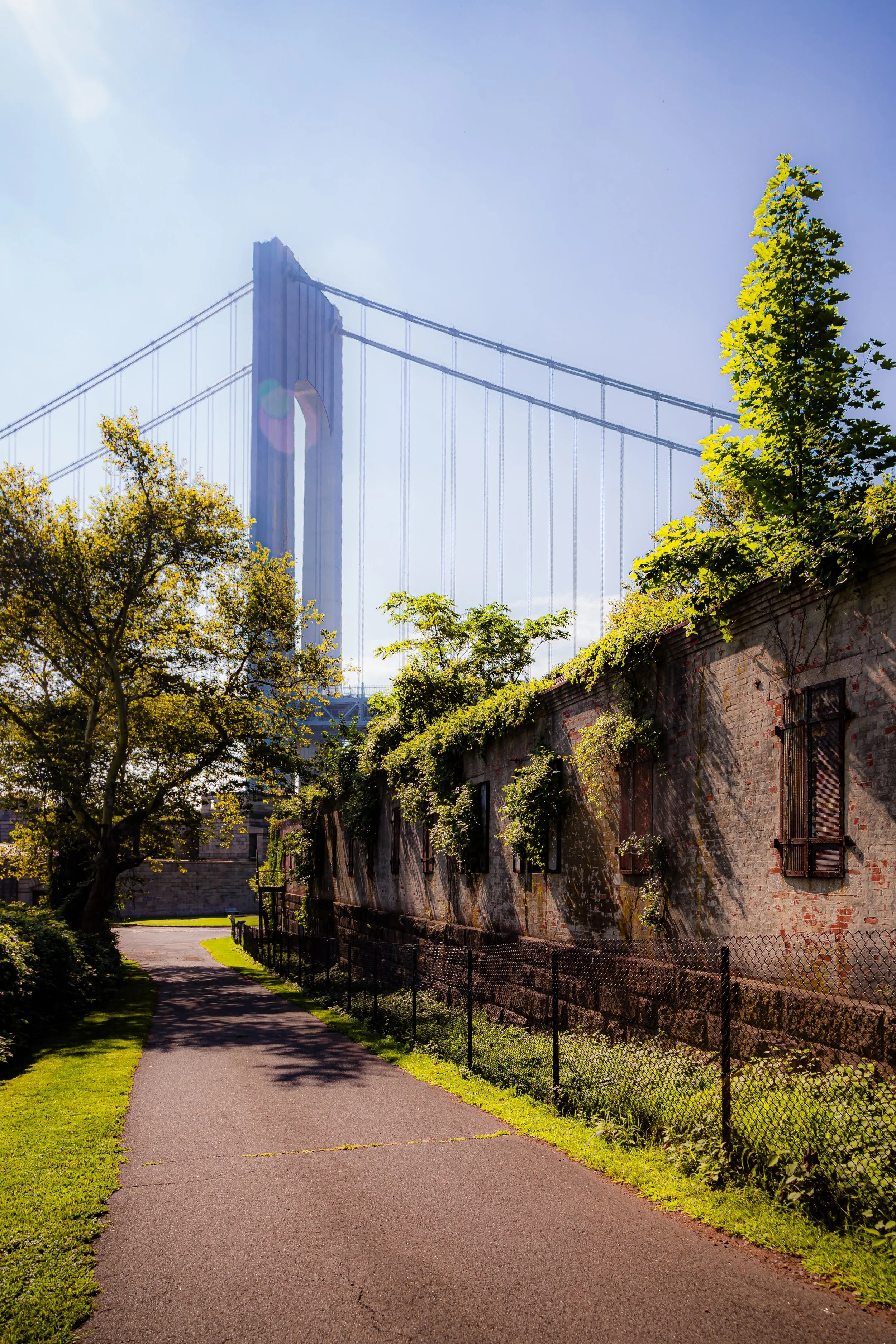 A pathway beside an old brick building with windows and vines, leading towards the New York City skyline and the Brooklyn Bridge.