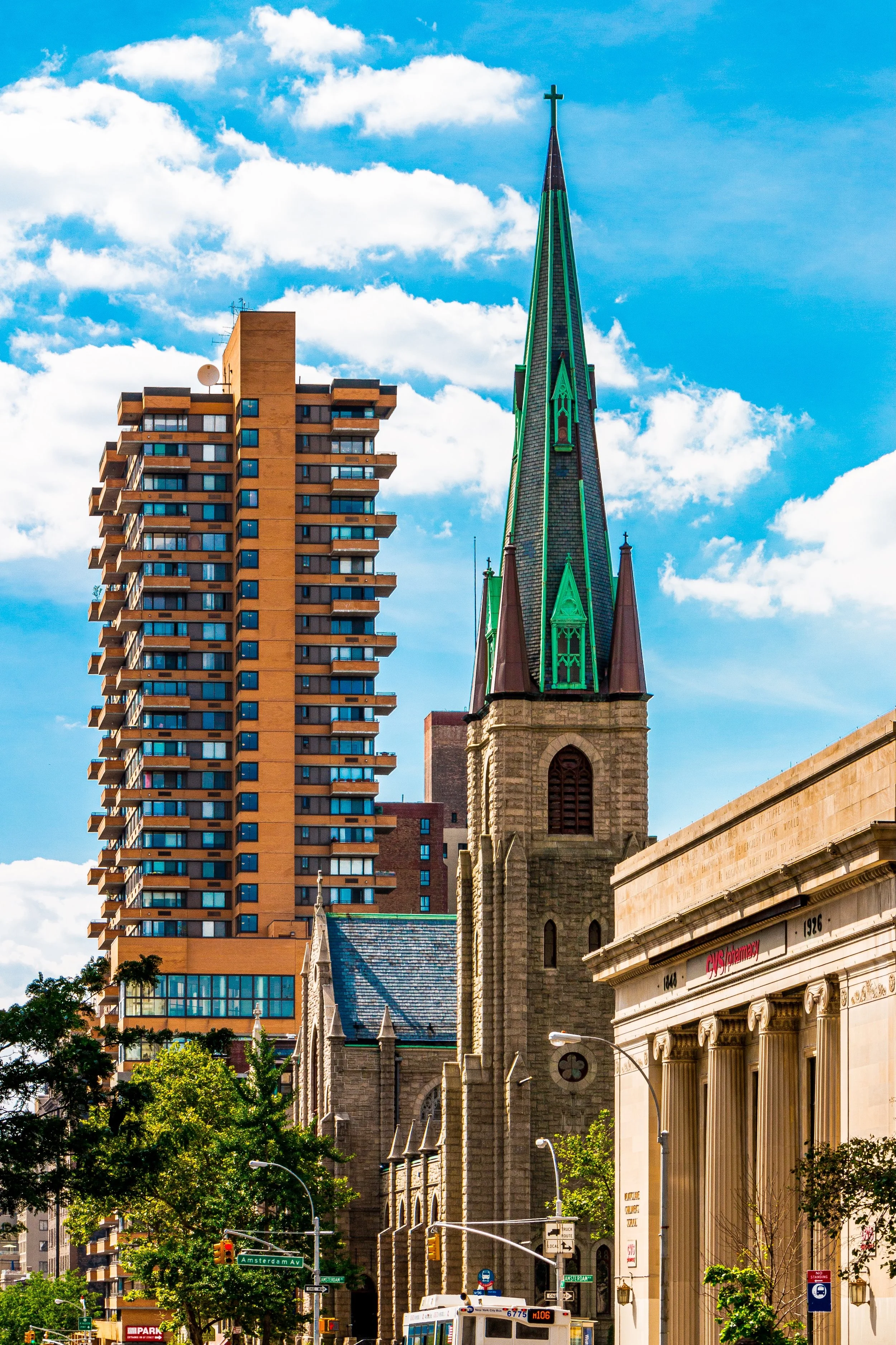 A city street scene with a church featuring a tall spire next to a modern high-rise apartment building, under a partly cloudy blue sky with trees and a USPS mailbox in the foreground.