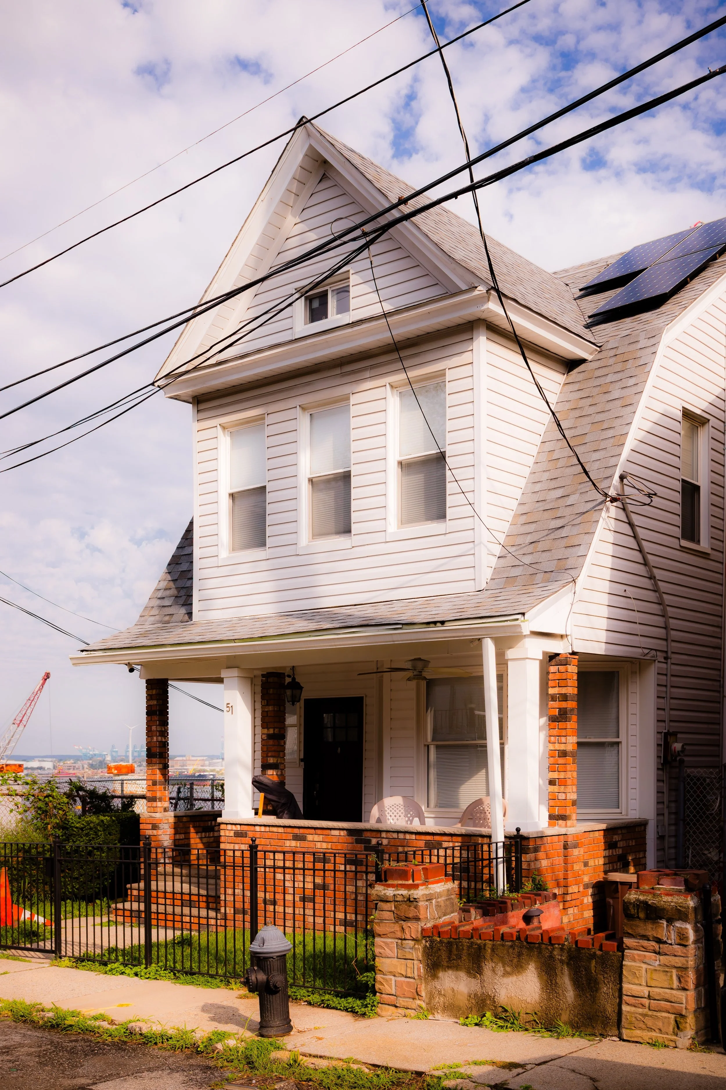 A two-story house with white siding, a front porch with brick pillars, black metal fence, and solar panels on the roof against a partly cloudy sky.