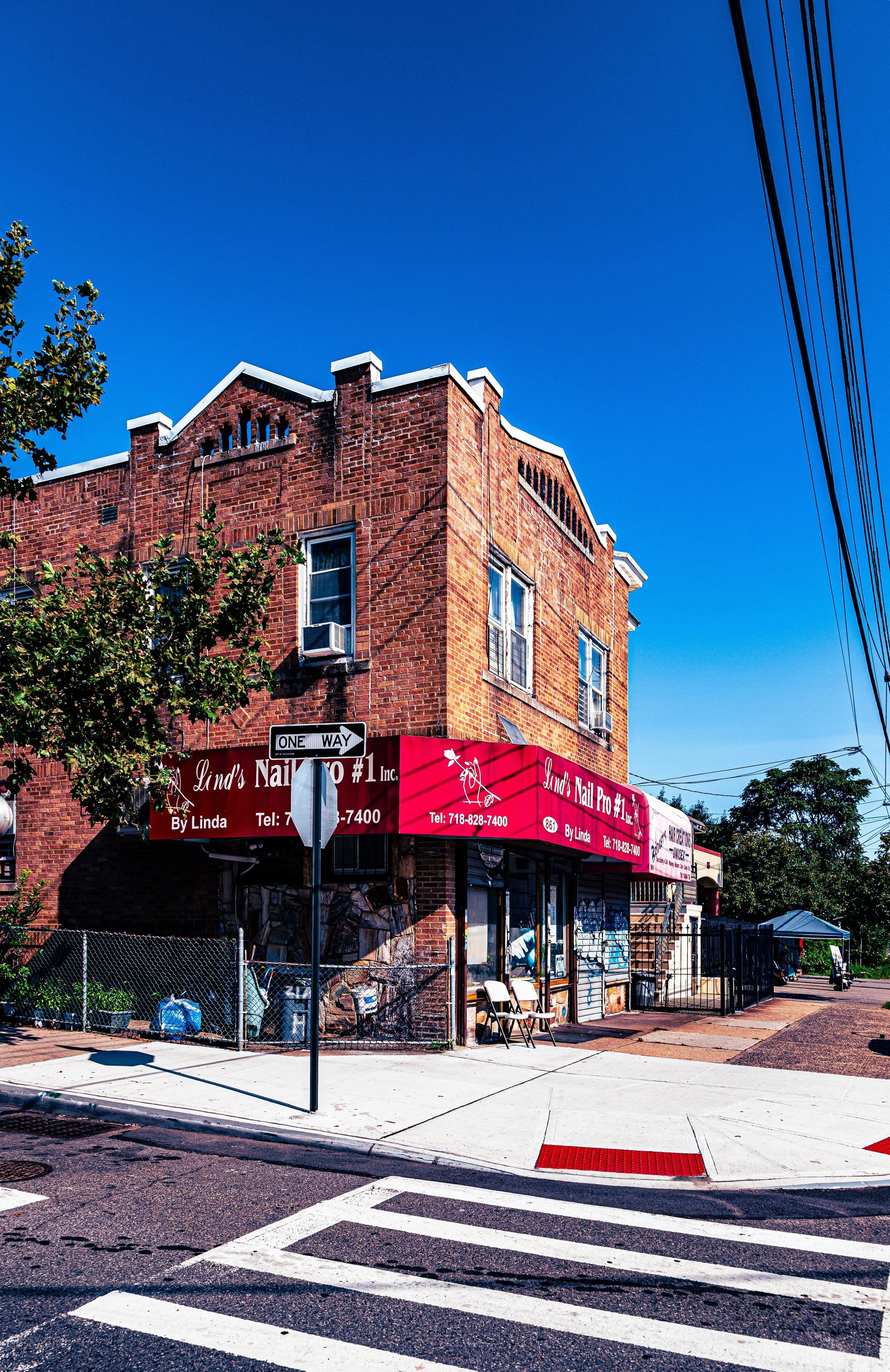 Brick building on a corner with a red sign for Linda's Nail Pro #1 Inc., with a one-way sign and crosswalk in the foreground under a clear blue sky.
