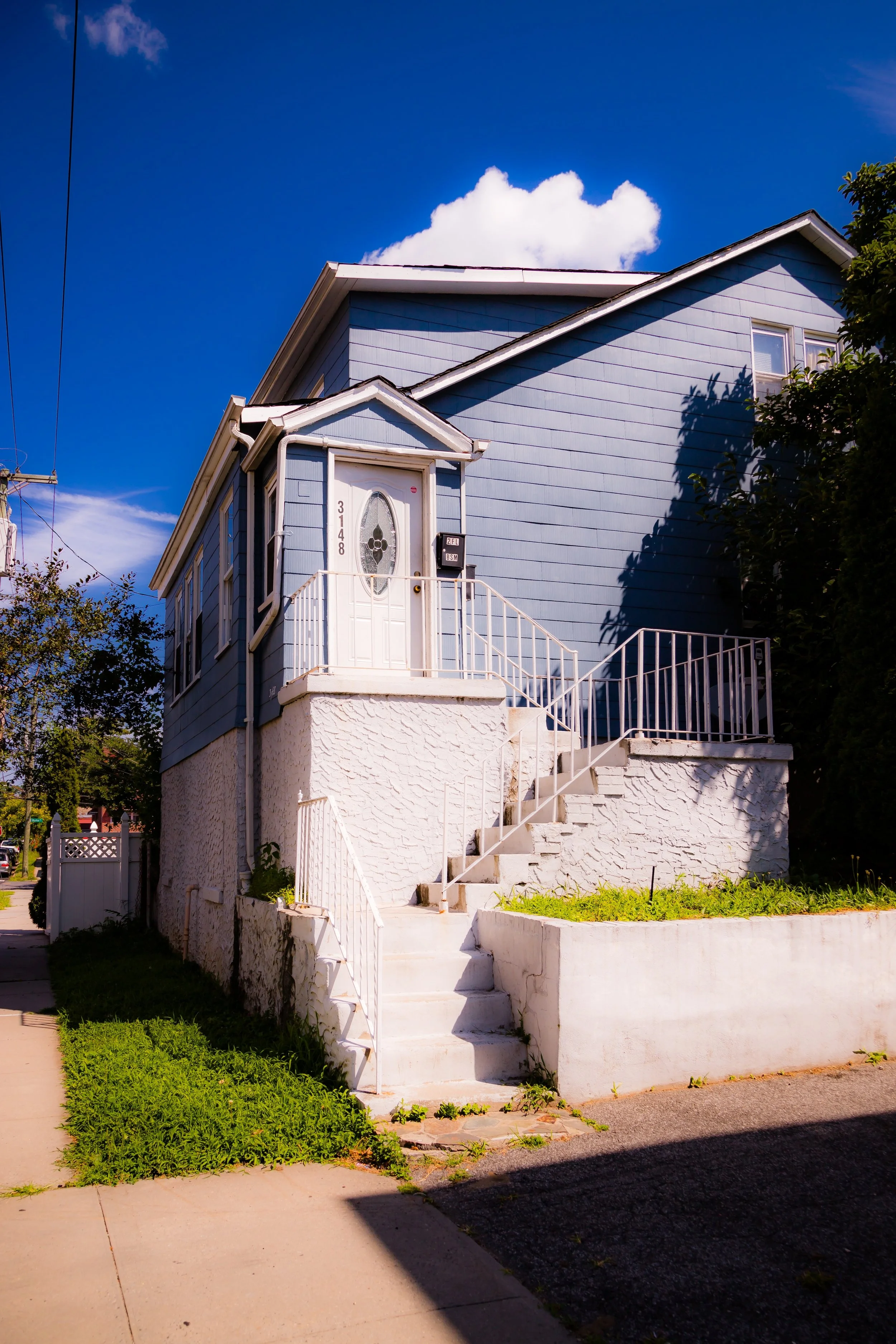 A two-story blue house with a white staircase leading up to the front door, situated on a sidewalk under a blue sky with a few clouds.
