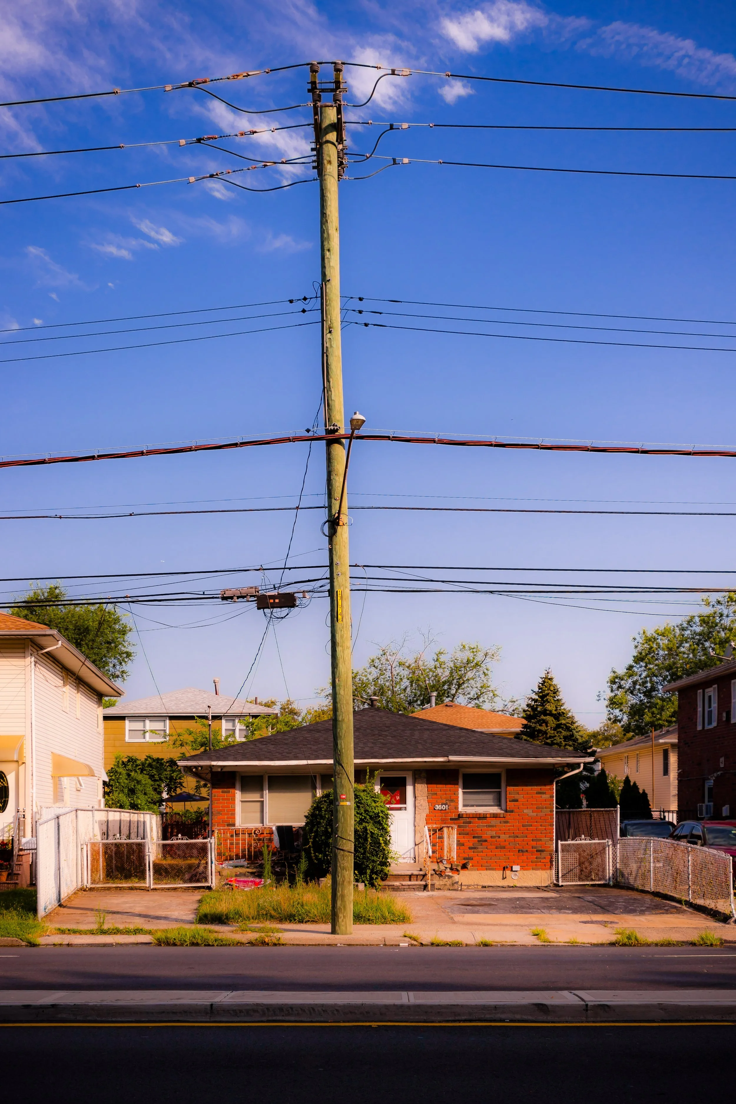 Residential house with a brick exterior, small front yard, and a driveway, with power lines and a utility pole in the foreground, under a partly cloudy blue sky.
