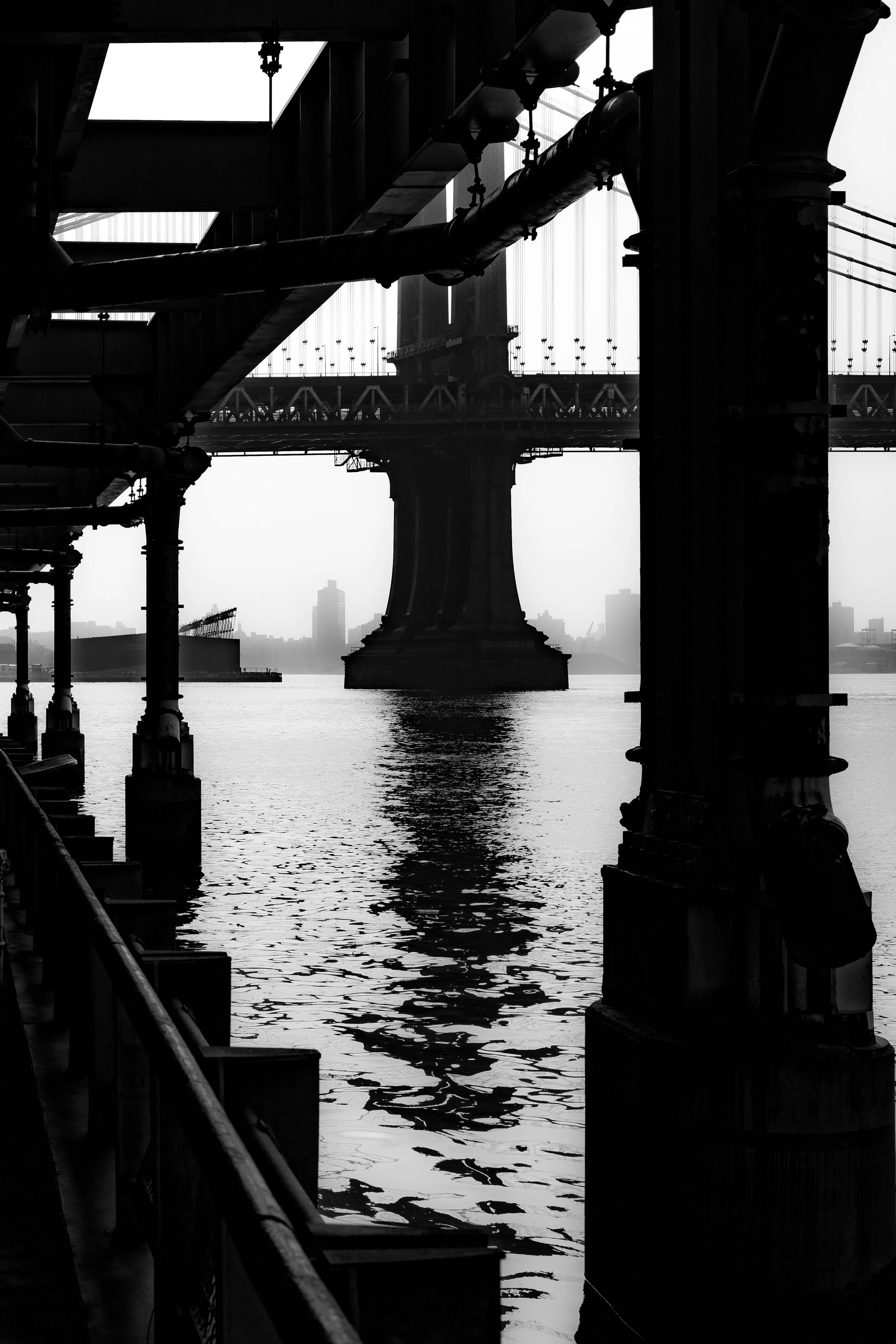 A black and white photo of a bridge seen from underneath with support beams, pipes, and a railing along the water."