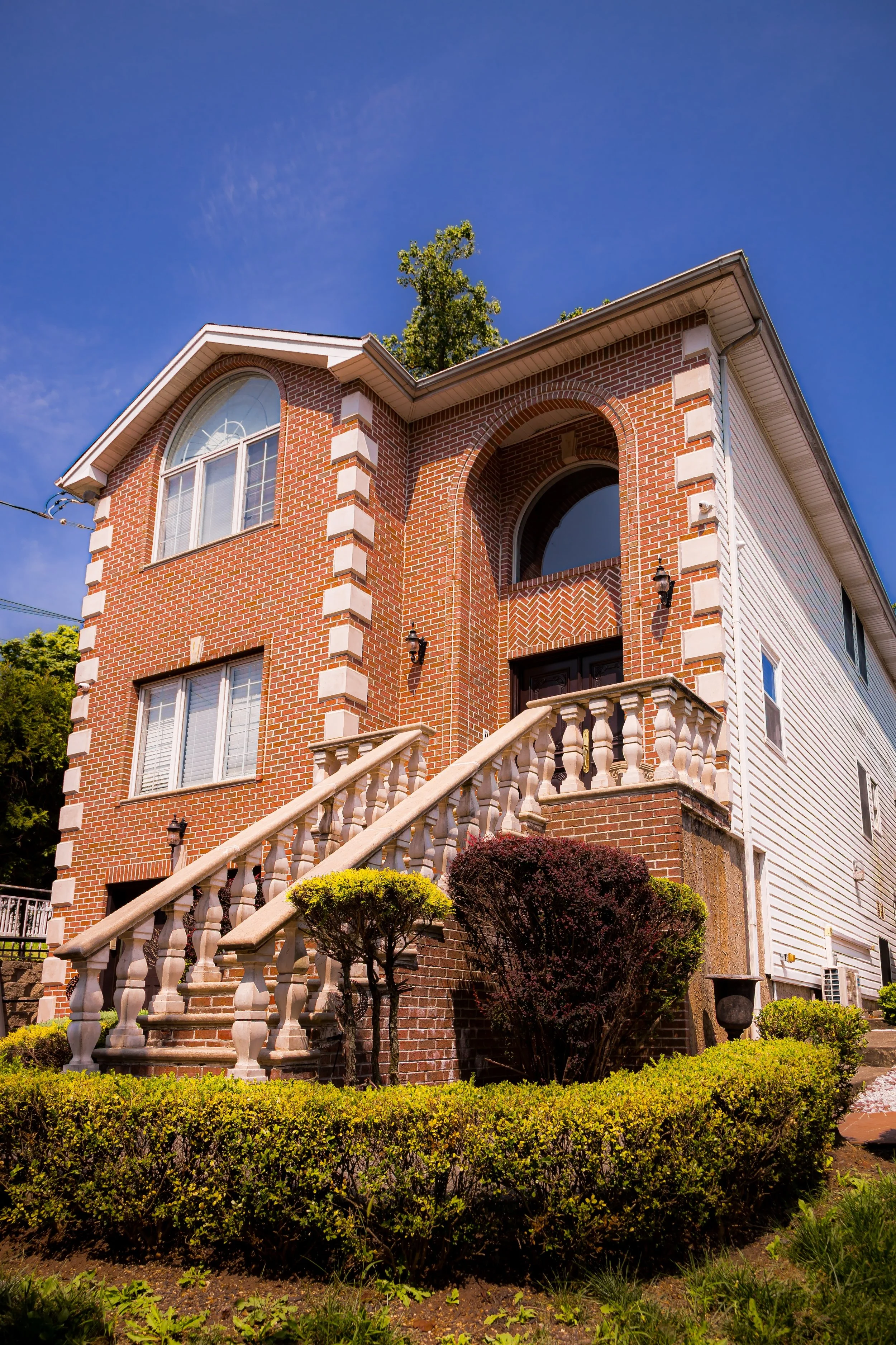 A multi-story brick house with a grand entrance, stairs, and a landscaped front yard under a clear blue sky.