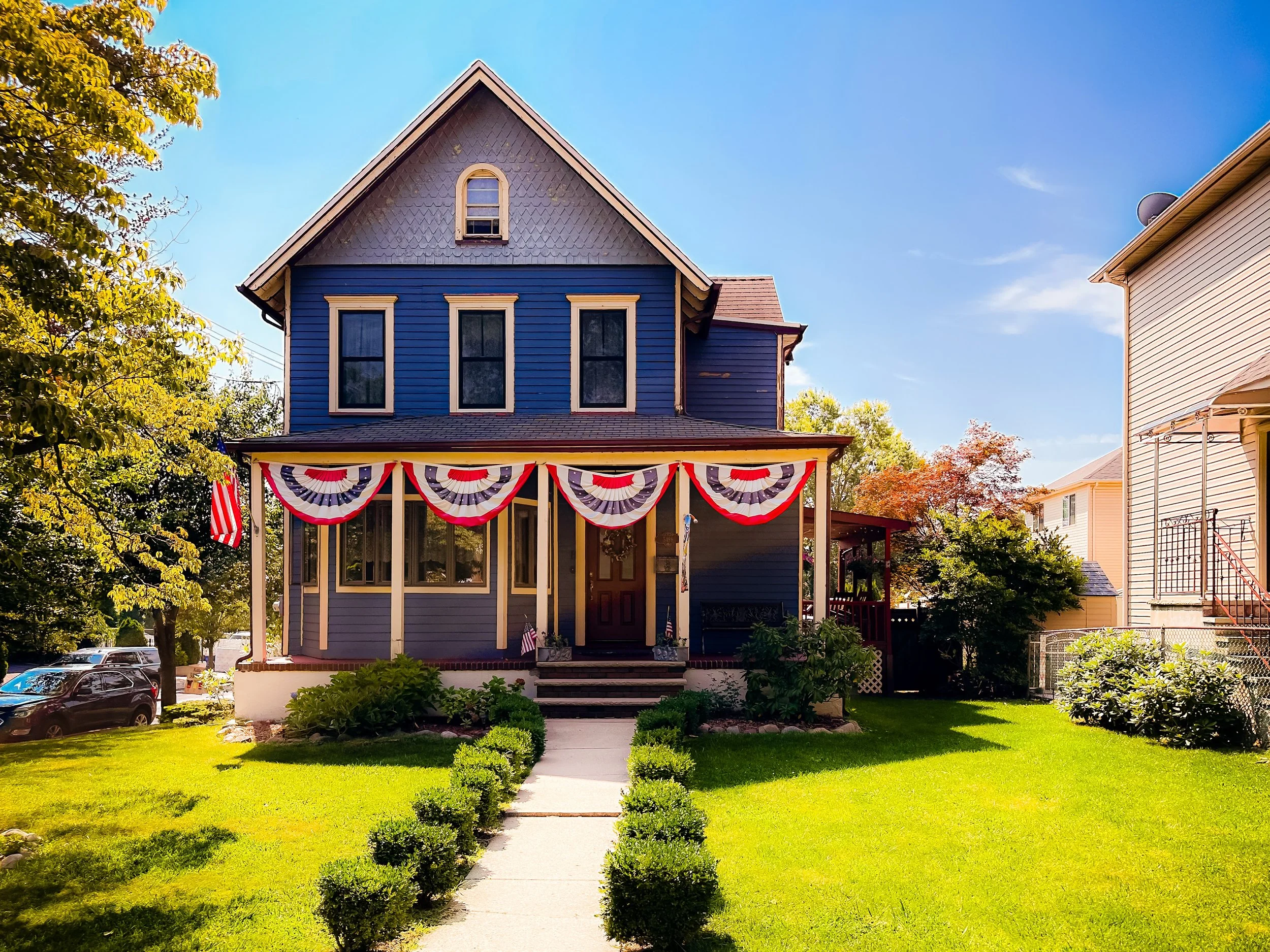 Blue house with patriotic decorations, including red, white, and blue bunting, and an American flag, on a sunny day with a green lawn and neighboring houses.