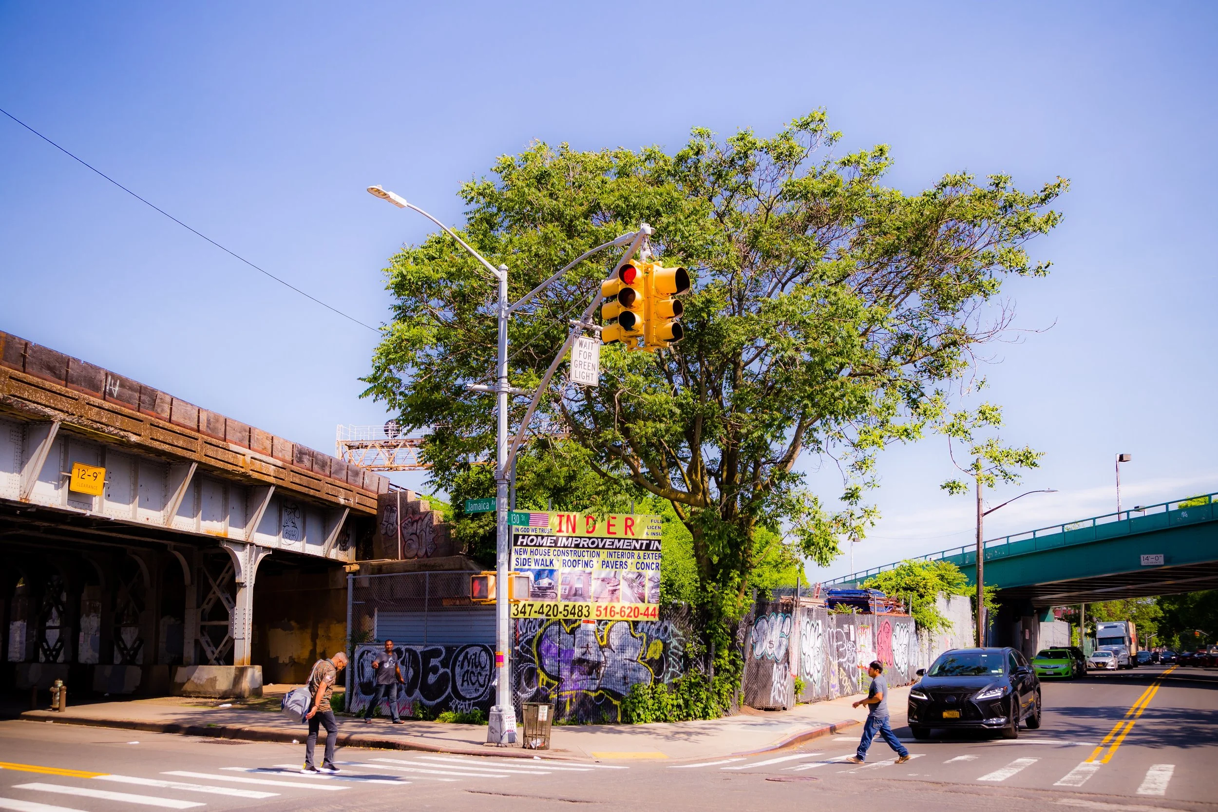 City street intersection with traffic light, people crossing, graffiti on walls, a large tree, an overpass, and vehicles.