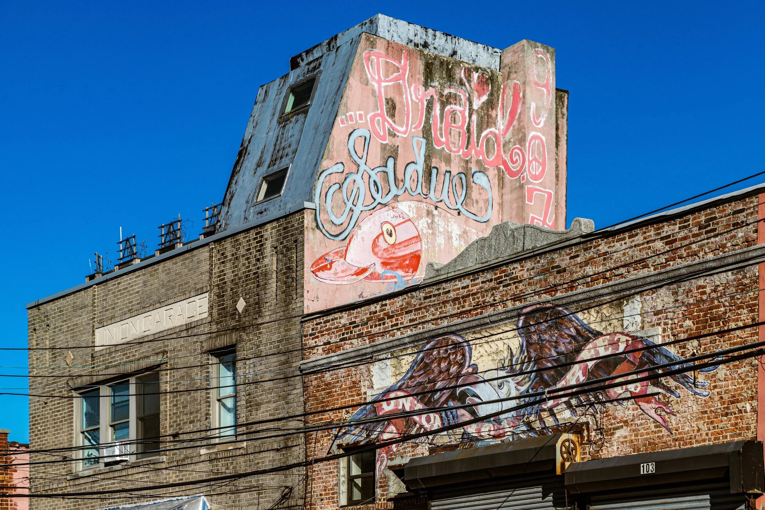 A brick building with street art murals; one mural features a red and white creature with large eyes, another depicts a lobster with wings and a banner. There is signage that says 'UNDERAGE' and house number 103.