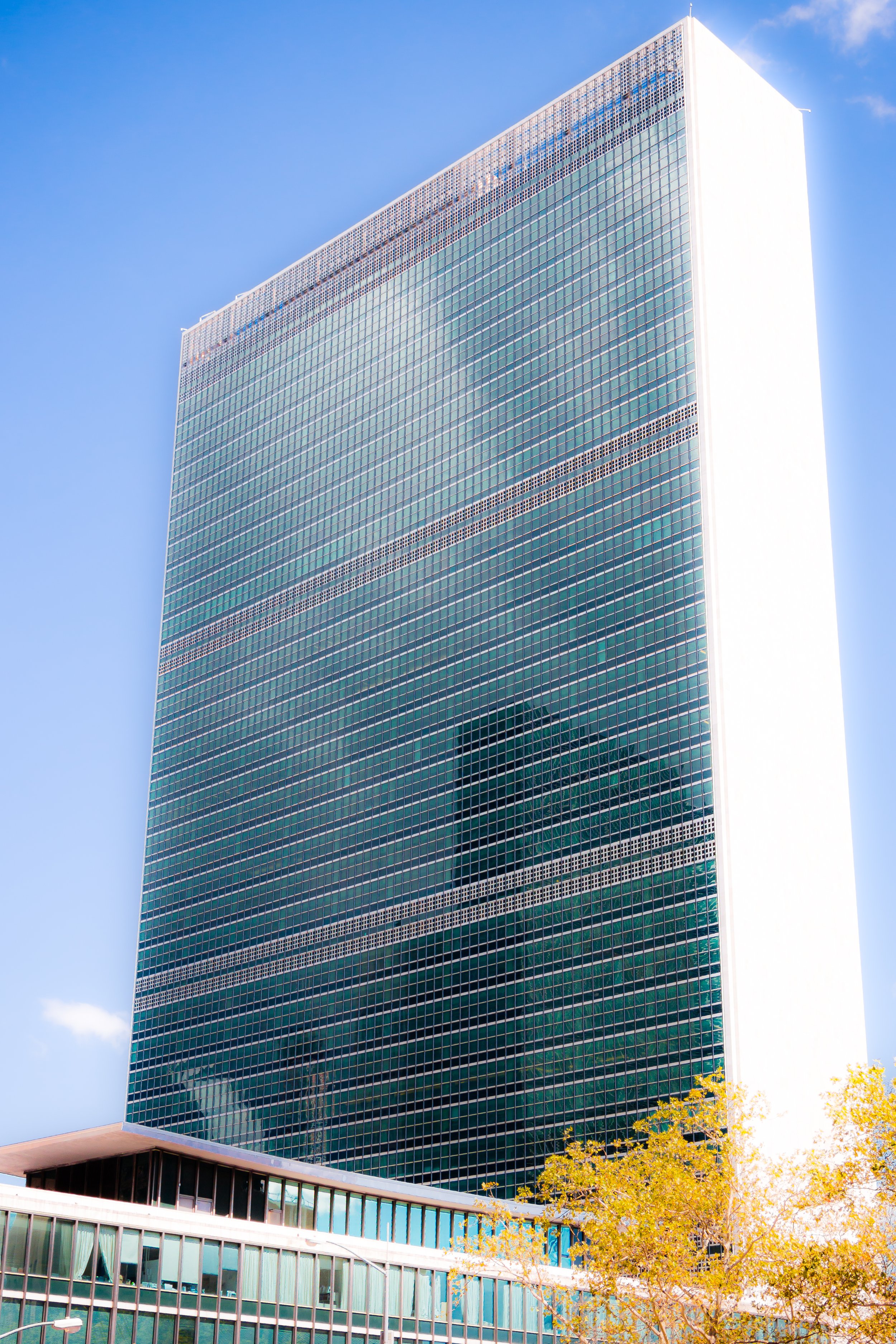 A tall modern glass skyscraper reflecting the blue sky and clouds, with some yellow autumn trees visible at the bottom.