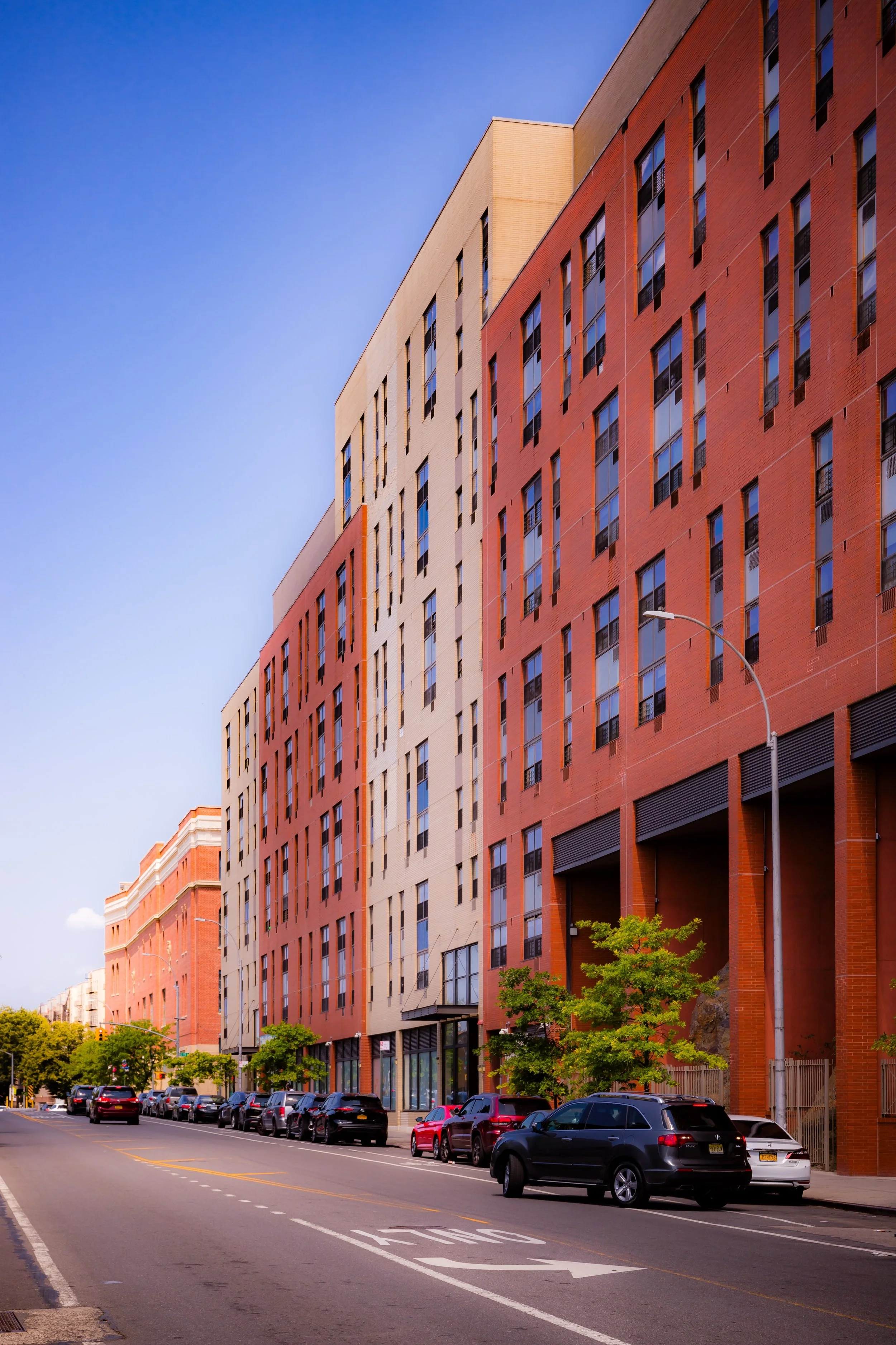 Street view of modern multi-story residential buildings with parked cars and trees, under a clear blue sky.
