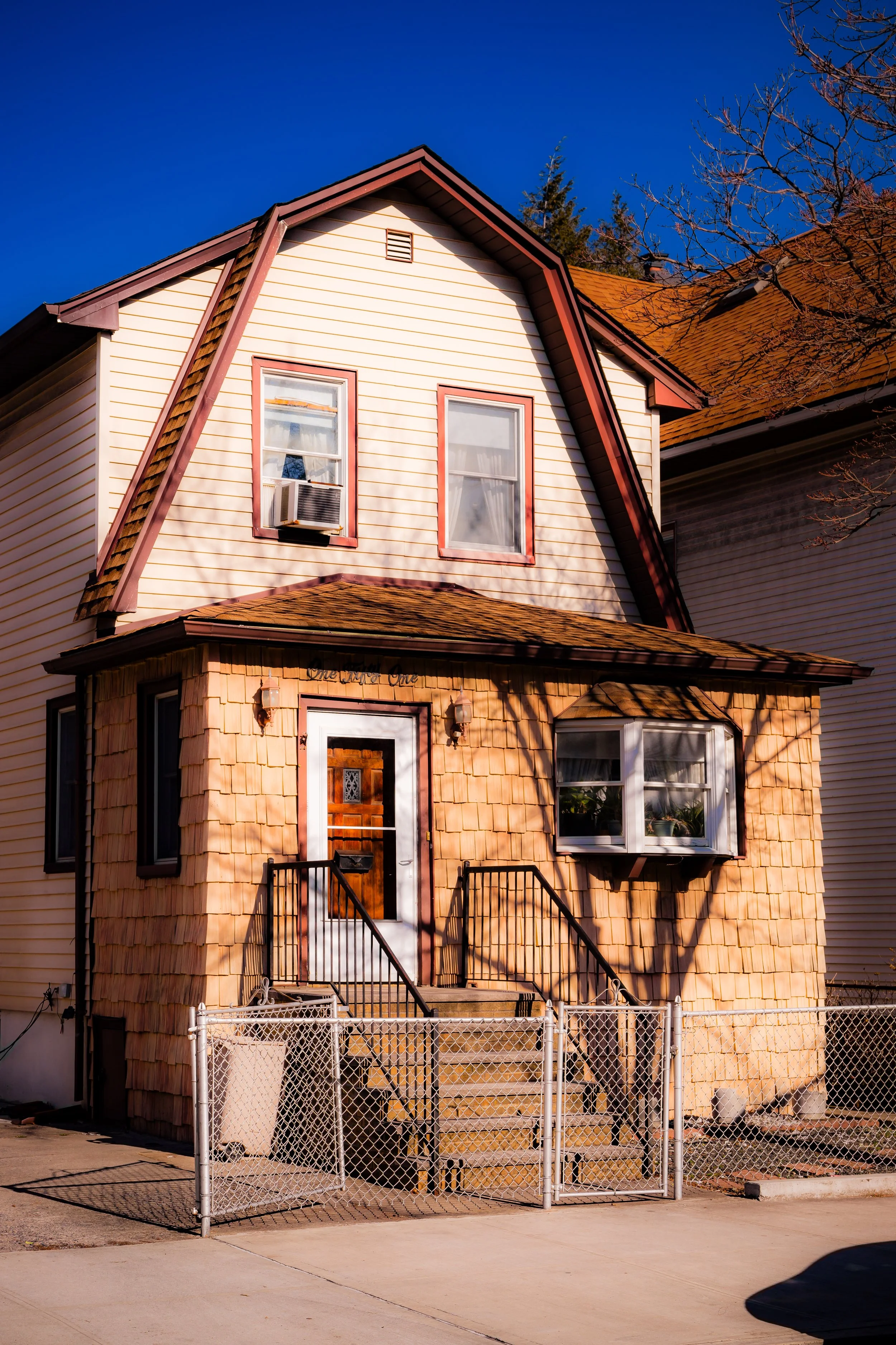 A two-story house with beige and brown siding, white-framed windows, and a small porch with stairs. The house has a decorative entryway with a wooden door and a small bay window. The sky is clear and blue.