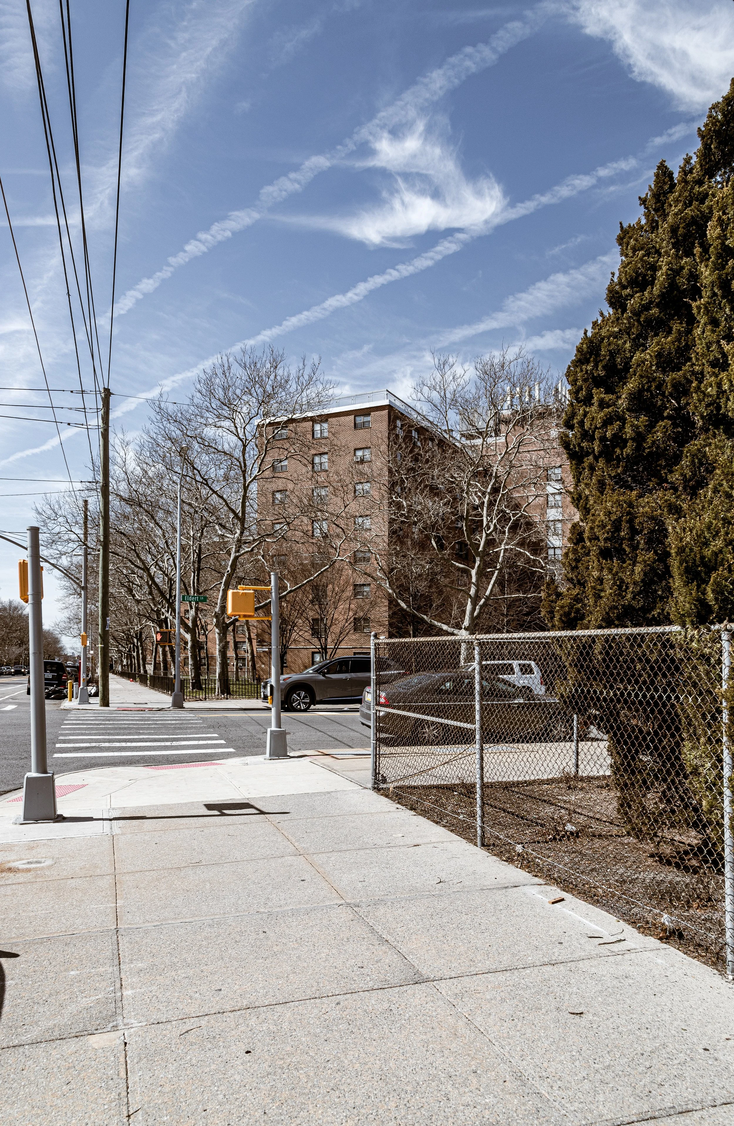 City street corner with a crosswalk, traffic lights, parked cars, leafless trees, a chain-link fence, and a multi-story brick building under a blue sky with clouds and contrails.