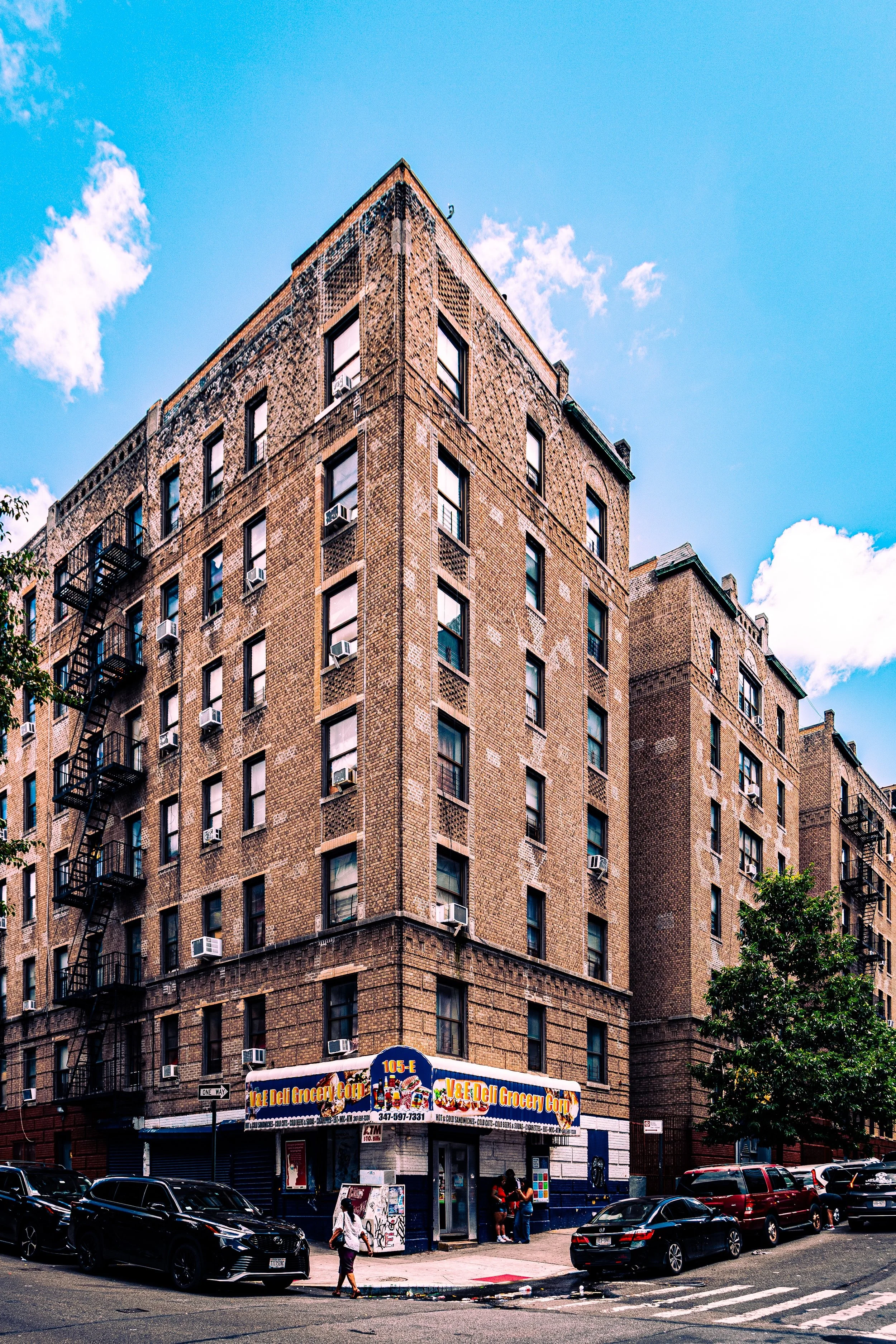 A multi-story brick apartment building with fire escapes and air conditioning units on the windows, situated at a street corner with parked cars and a small grocery store at the entrance. There are a few people walking and standing outside the store under a colorful awning. The sky is mostly clear with some clouds.