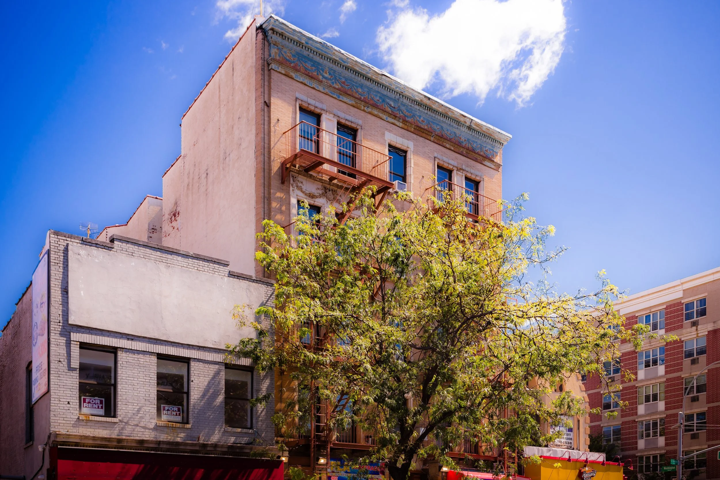 Multi-story buildings with a tree in front, some windows with 'For Rent' signs, and a bright blue sky.
