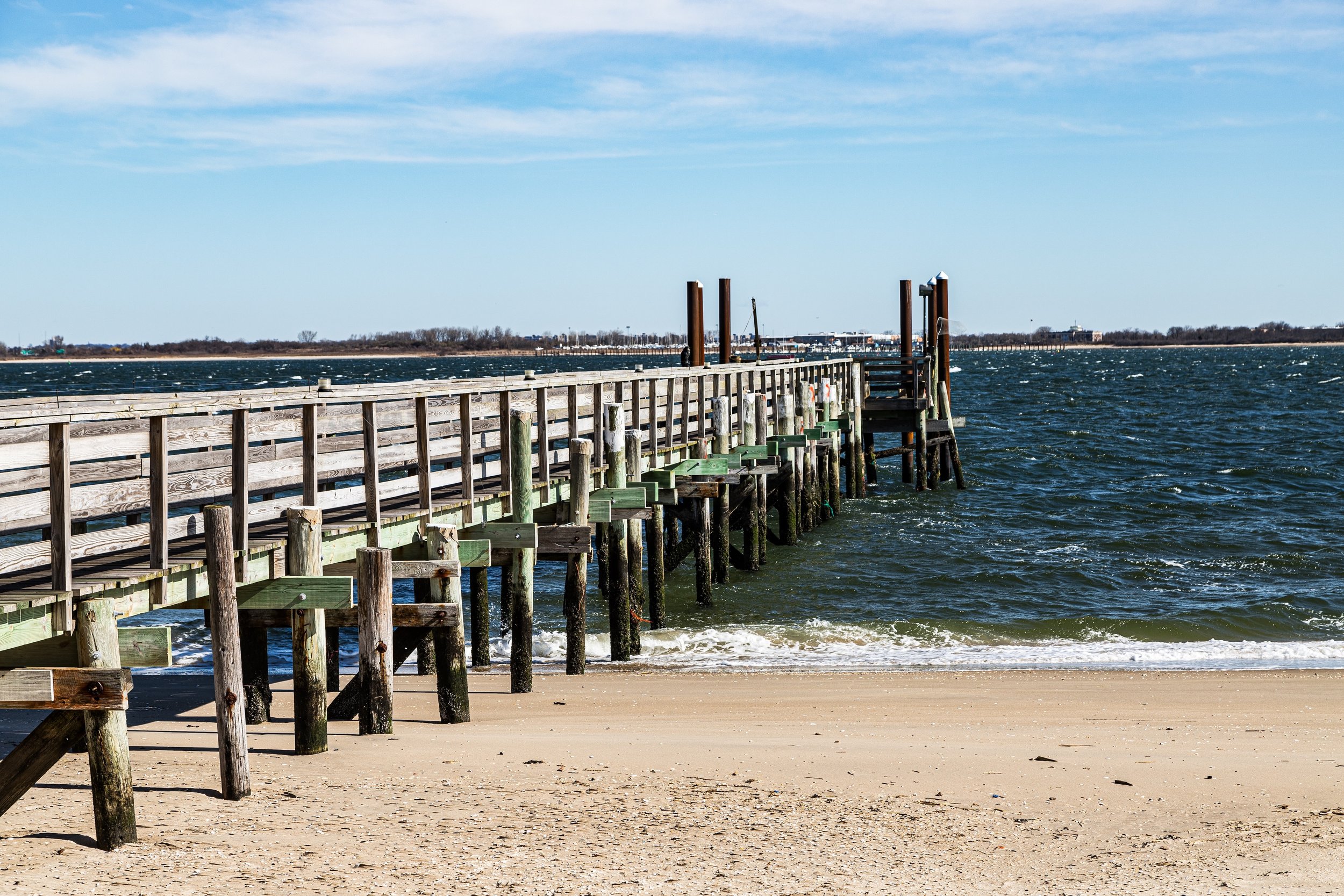 A wooden pier extending into the water at a beach, with sand in the foreground and blue sky above.