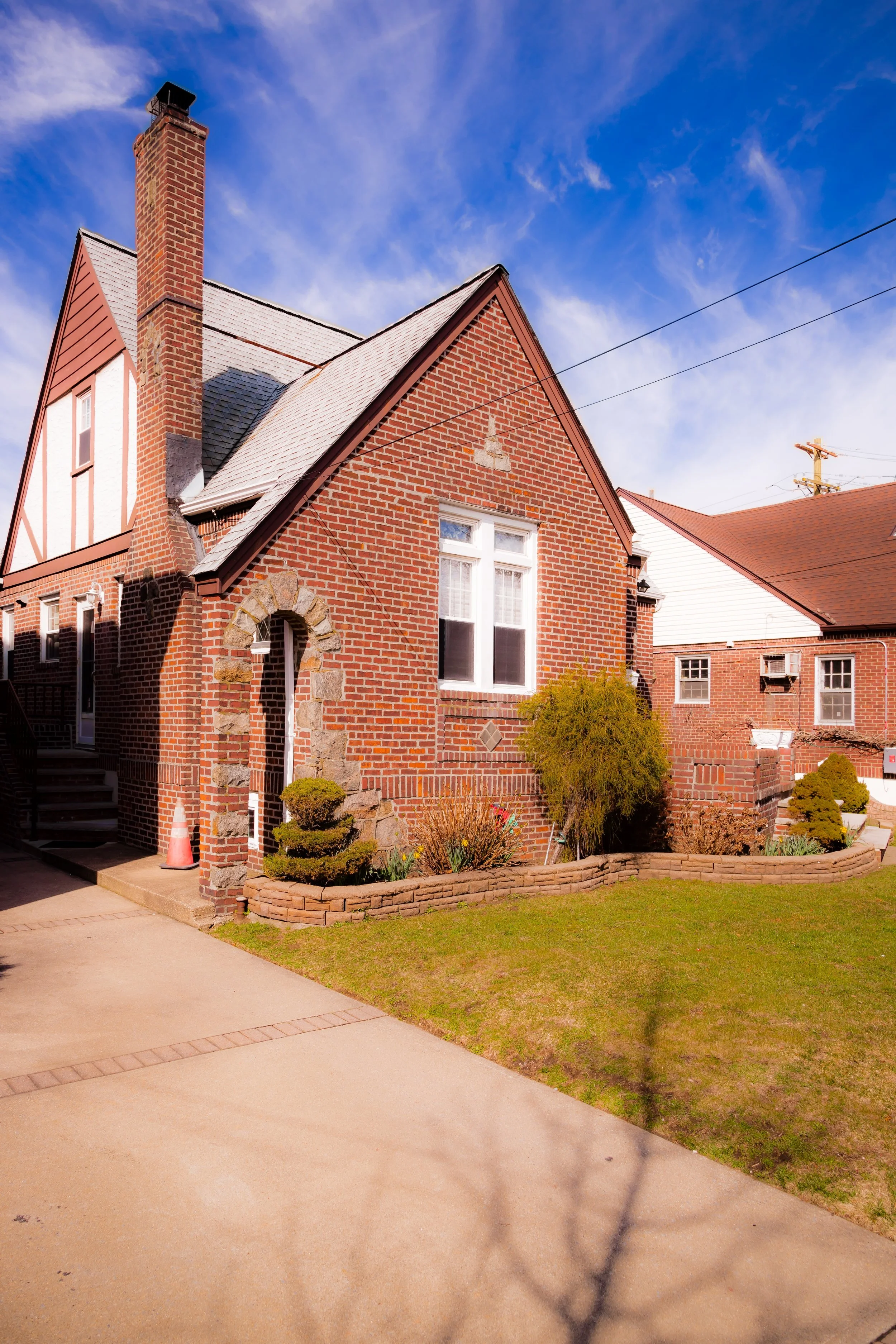 A brick house with a chimney and a gabled roof, front yard with shrubs and plants, and a sidewalk in front.