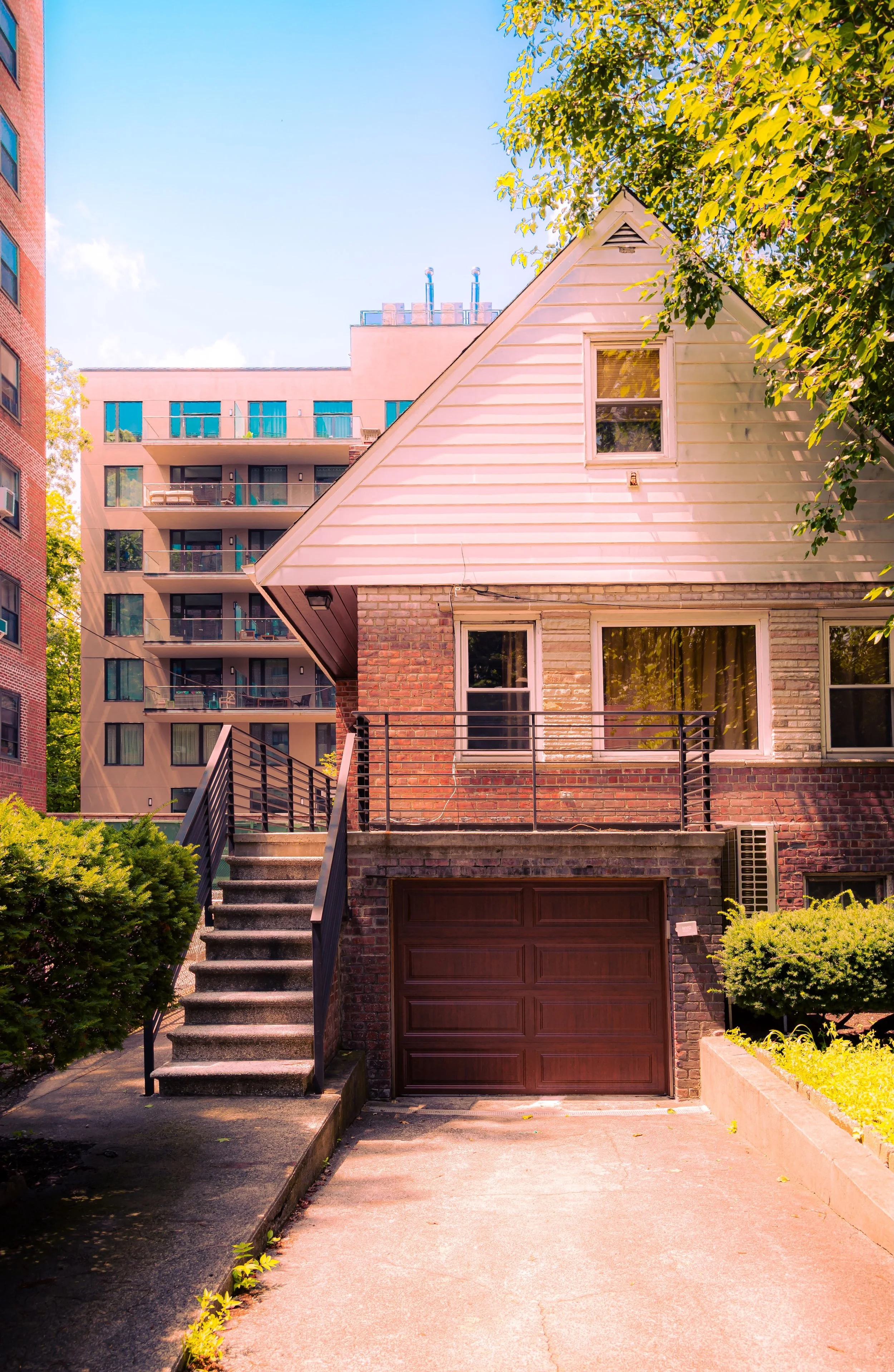 A brick and white house with a staircase leading up to a balcony and a garage door, surrounded by green bushes and trees, with a large modern apartment building in the background.