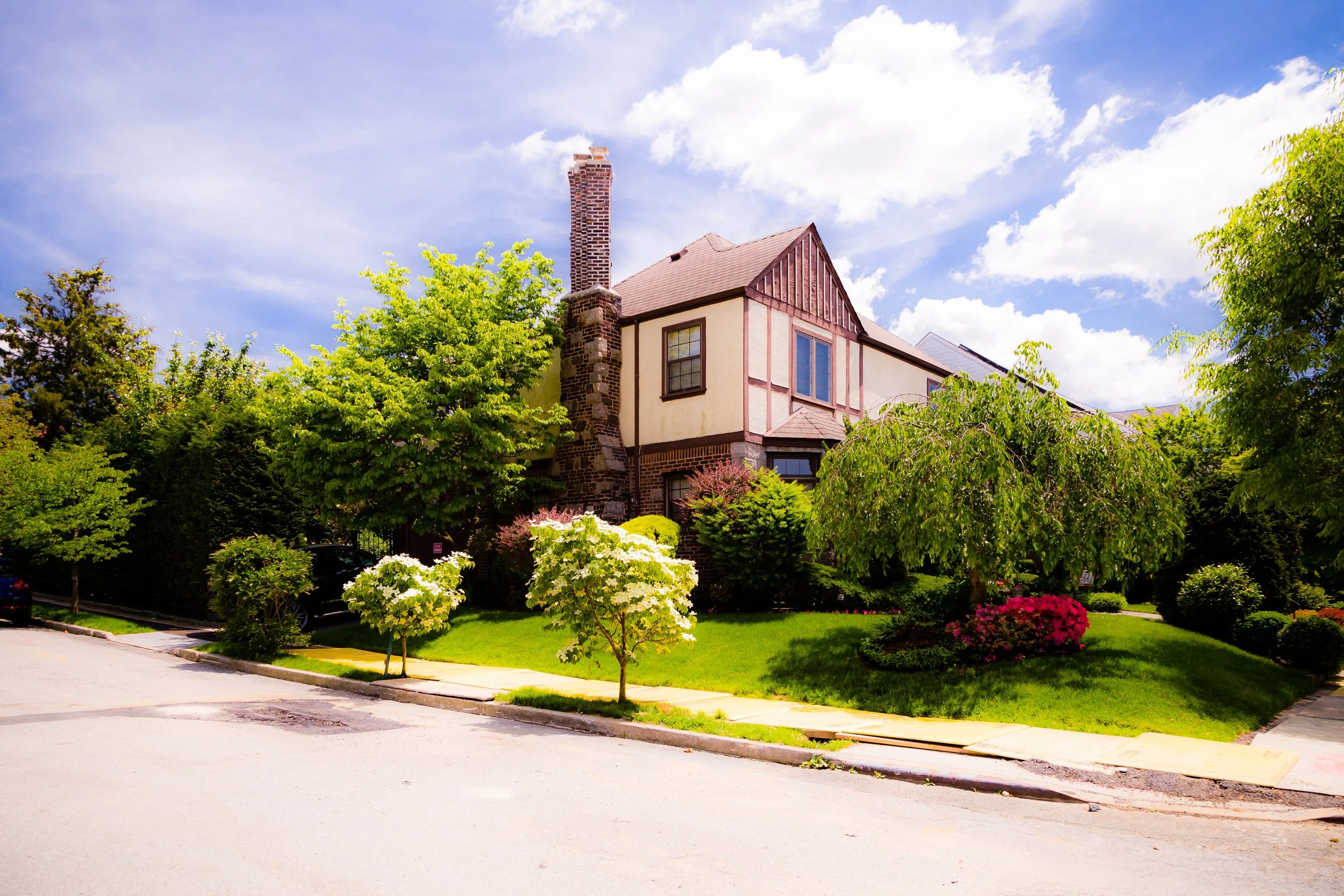 A house with a mix of stone and siding exterior, surrounded by a lush garden with trees, bushes, and flowers, under a partly cloudy sky.