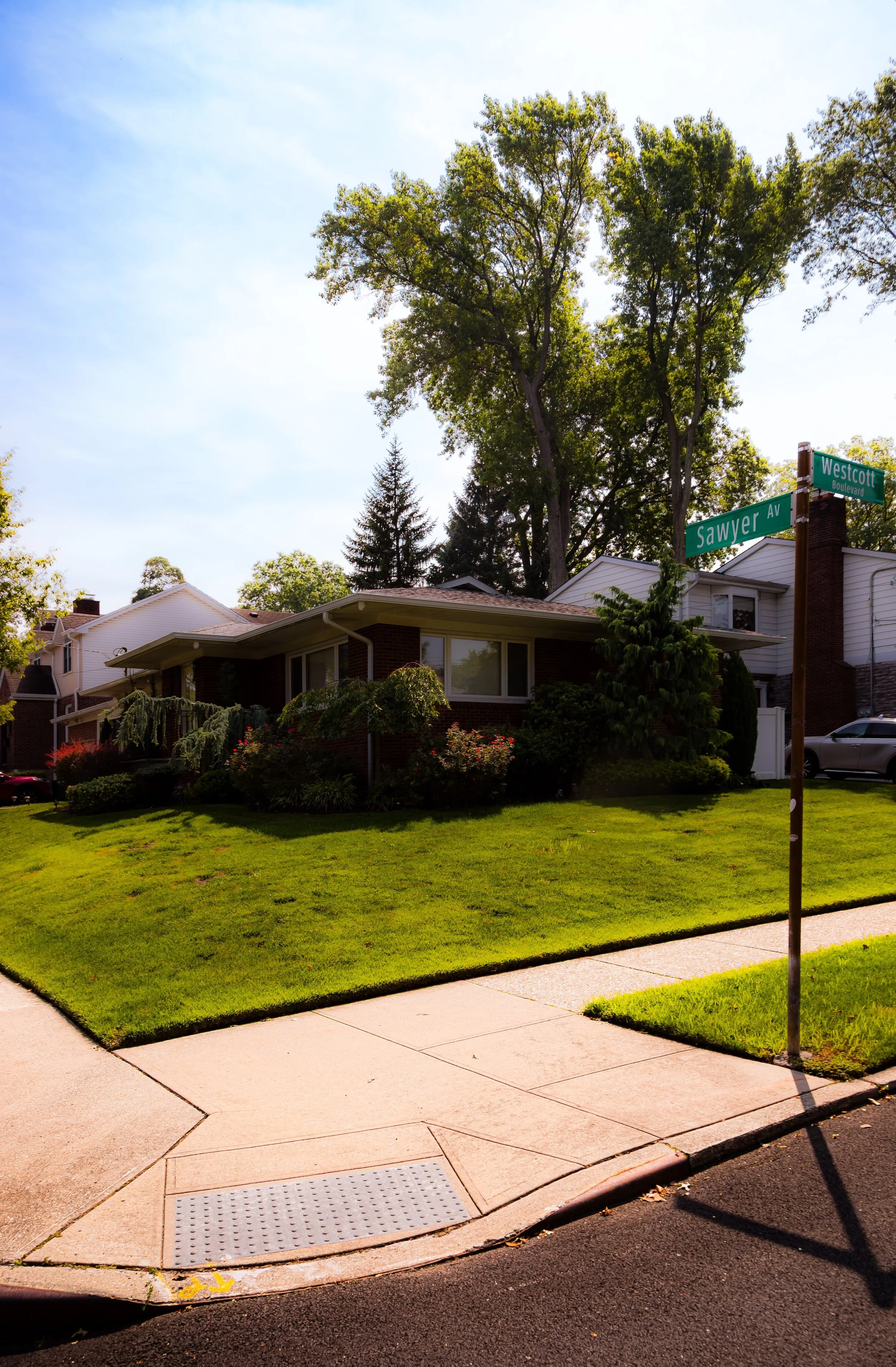 Street corner with a house, manicured lawn, trees, and a street sign showing Westcott Boulevard and Sawyer Avenue.