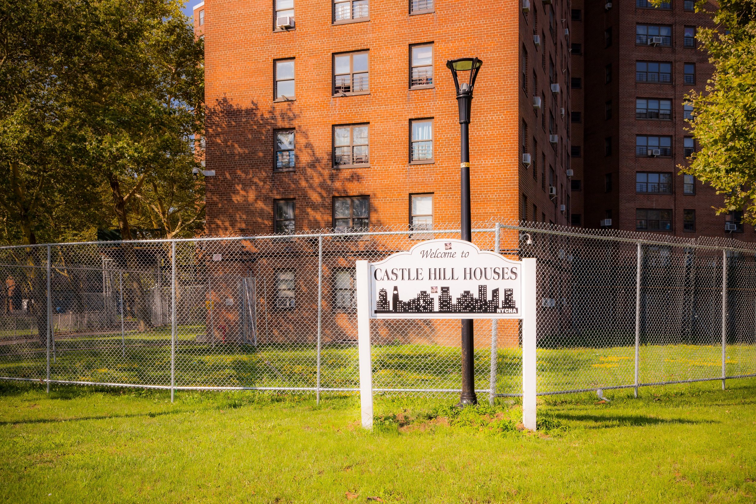 A fenced outdoor area with a sign that reads 'Welcome to Castle Hill Houses' and features a silhouette of a city skyline. Behind the fence is a grassy area and a large brick apartment building, with trees and a streetlamp in the foreground.