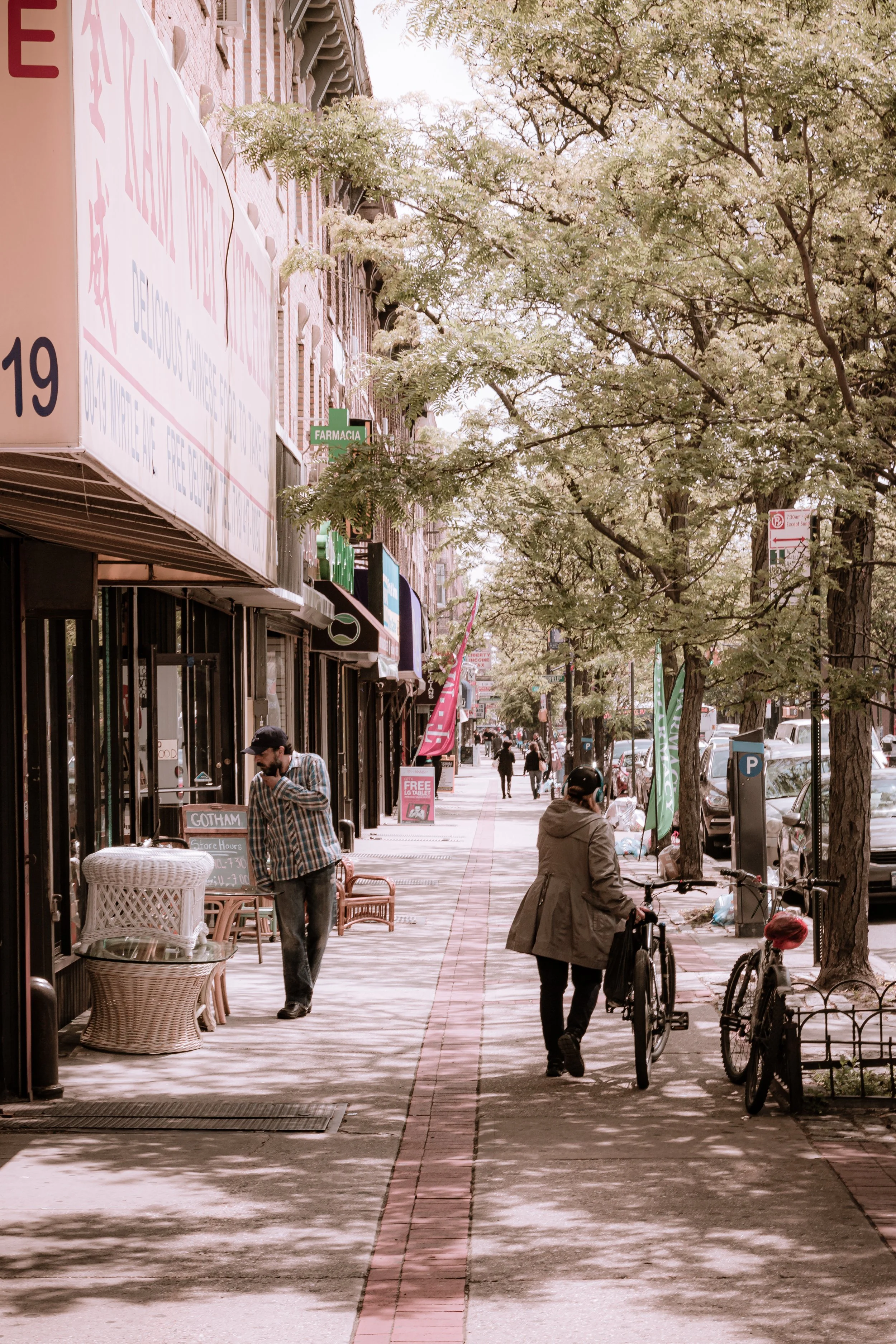 City sidewalk with people walking and biking, storefronts on the left, trees lining the street, parked cars on the right, and signs including a pharmacy sign.