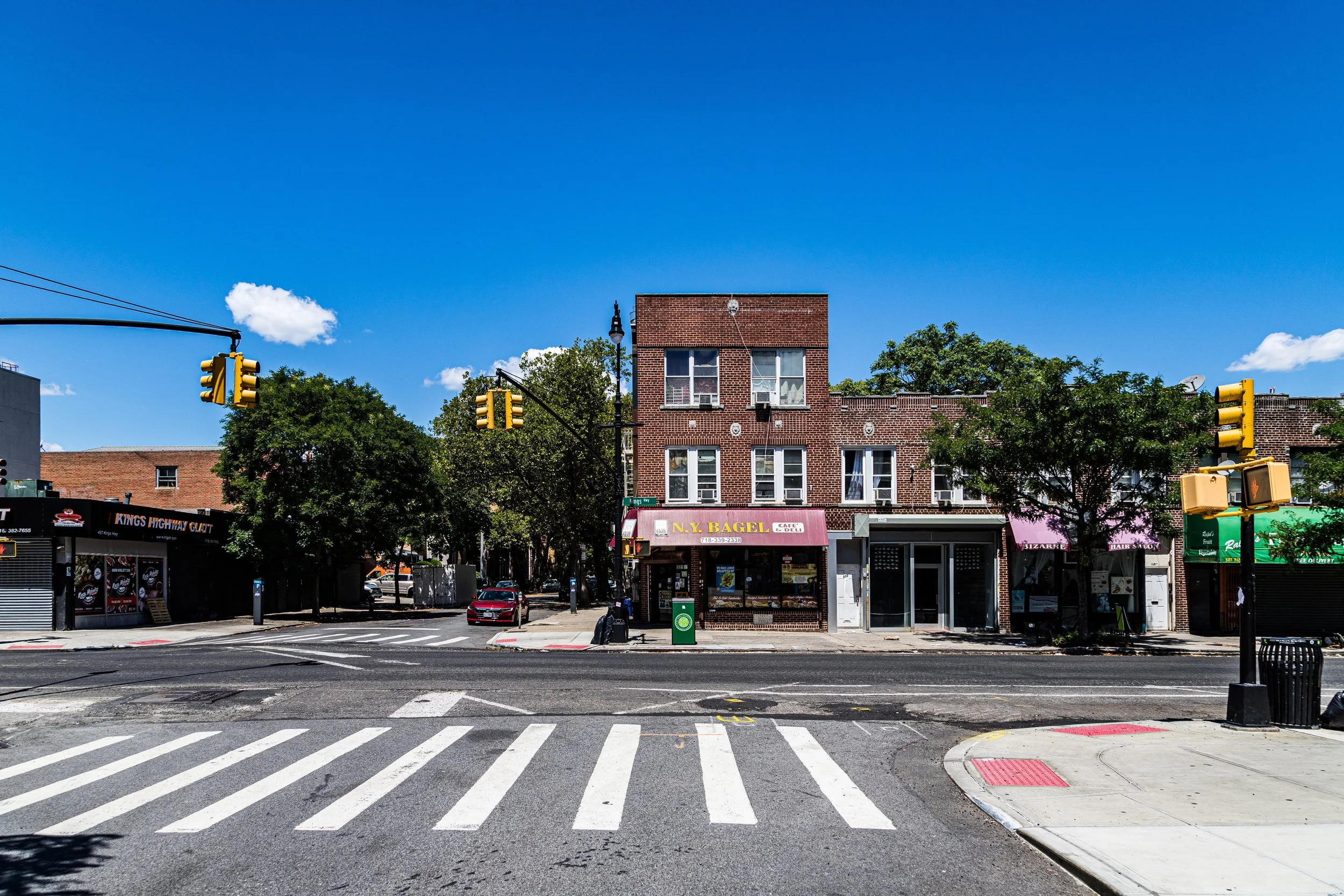 Street view of a crosswalk in front of small storefronts, including a bagel shop, with trees and traffic lights against a blue sky.