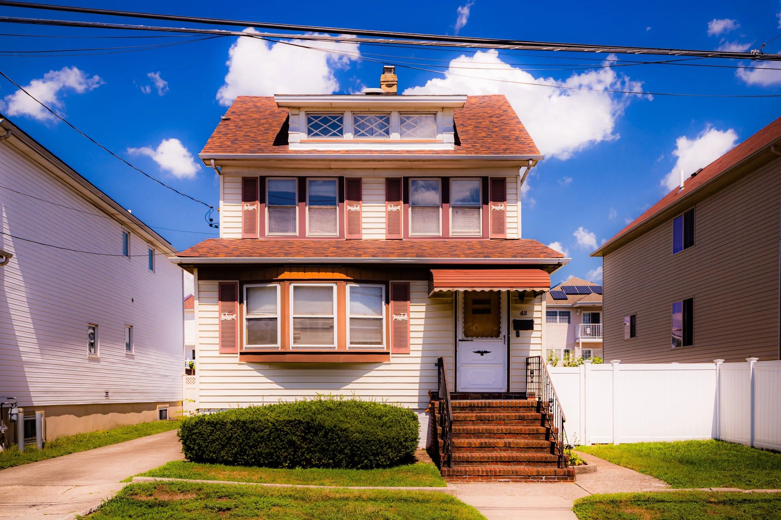 A three-story house with beige siding, brown roof, and a small front yard with a bush. Front steps lead to a white door with the house number 49, and there are five windows on the front, some with red shutters. The sky is blue with white clouds.