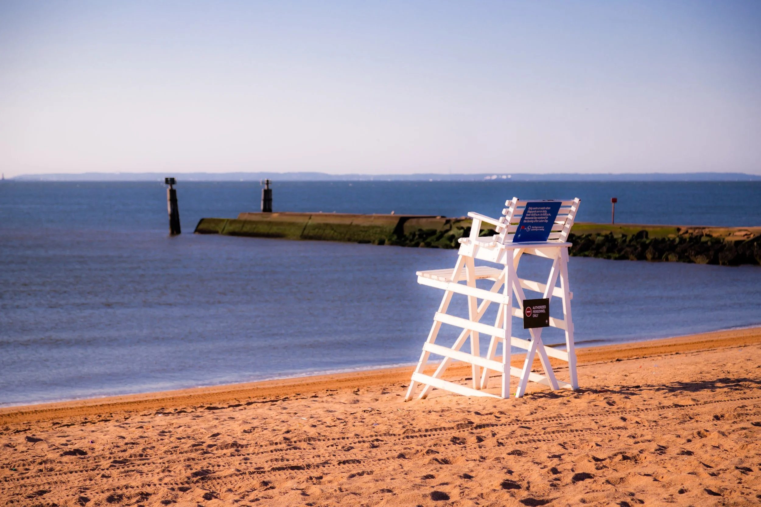 A white lifeguard stand on a sandy beach near the water, with a sign on top, and a sign indicating 'Authorized Personnel Only' attached below. In the background, there is a small breakwater extending into the water with two posts and a red flag.