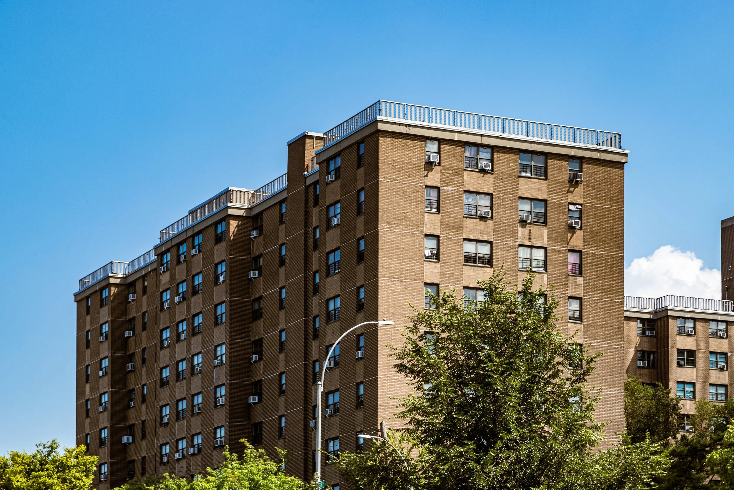 A tall brick apartment building with multiple windows and air conditioning units, surrounded by trees and a street lamp, under a clear blue sky.